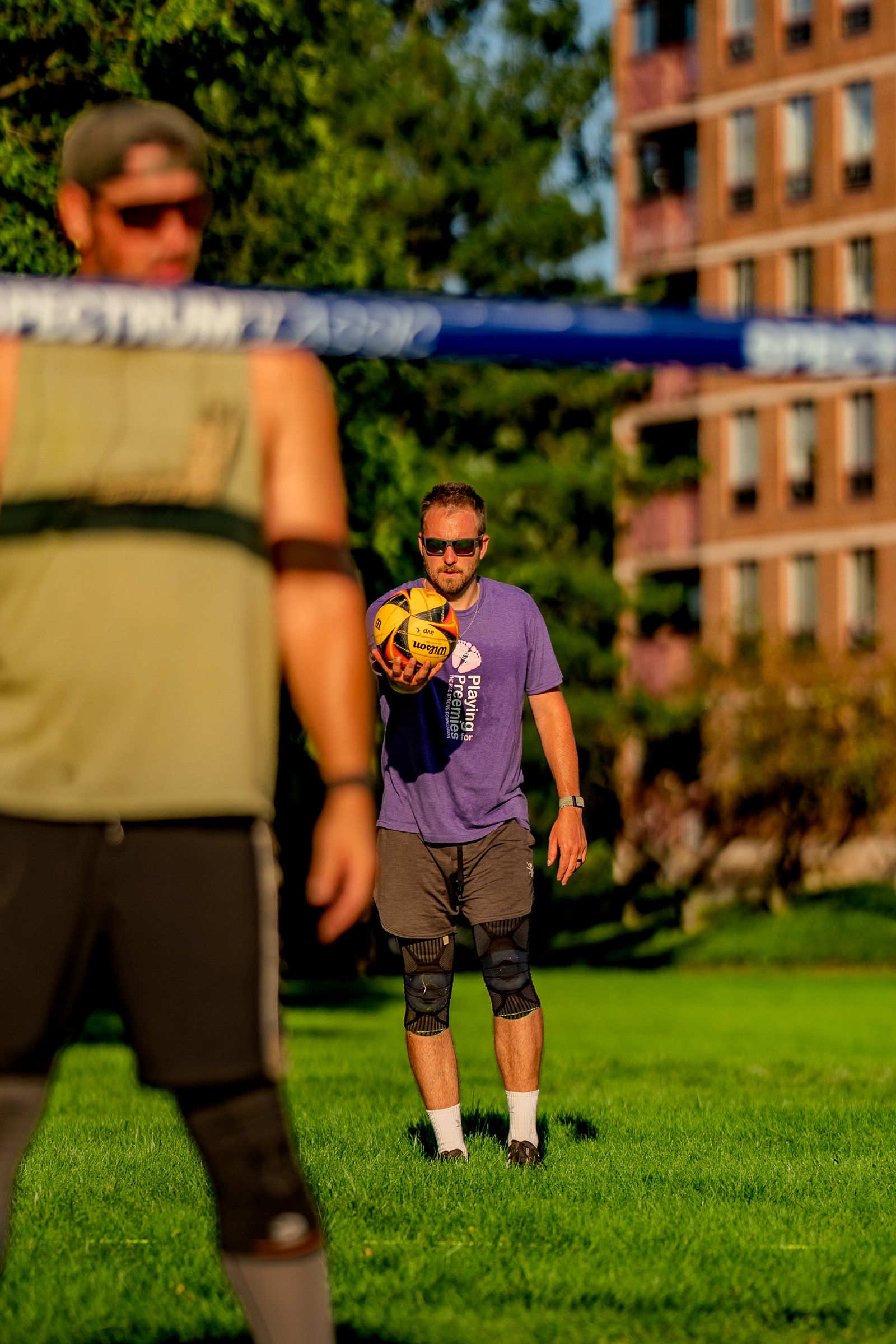 A player in a purple shirt holds a volleyball on a grassy court with a building in the background.