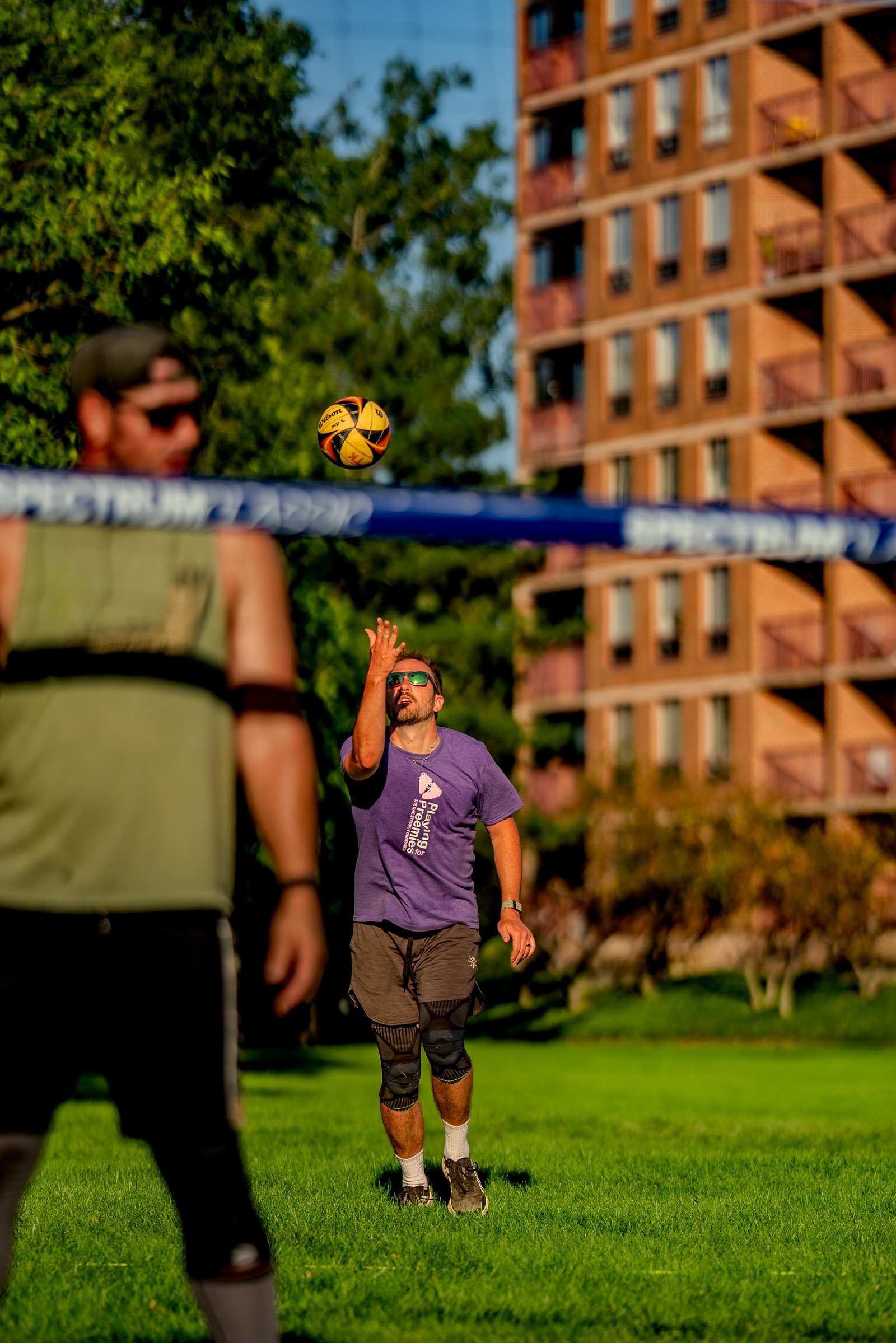 A player in a purple shirt sets the ball over the net during a volleyball game, with another player in a green shirt in the foreground.