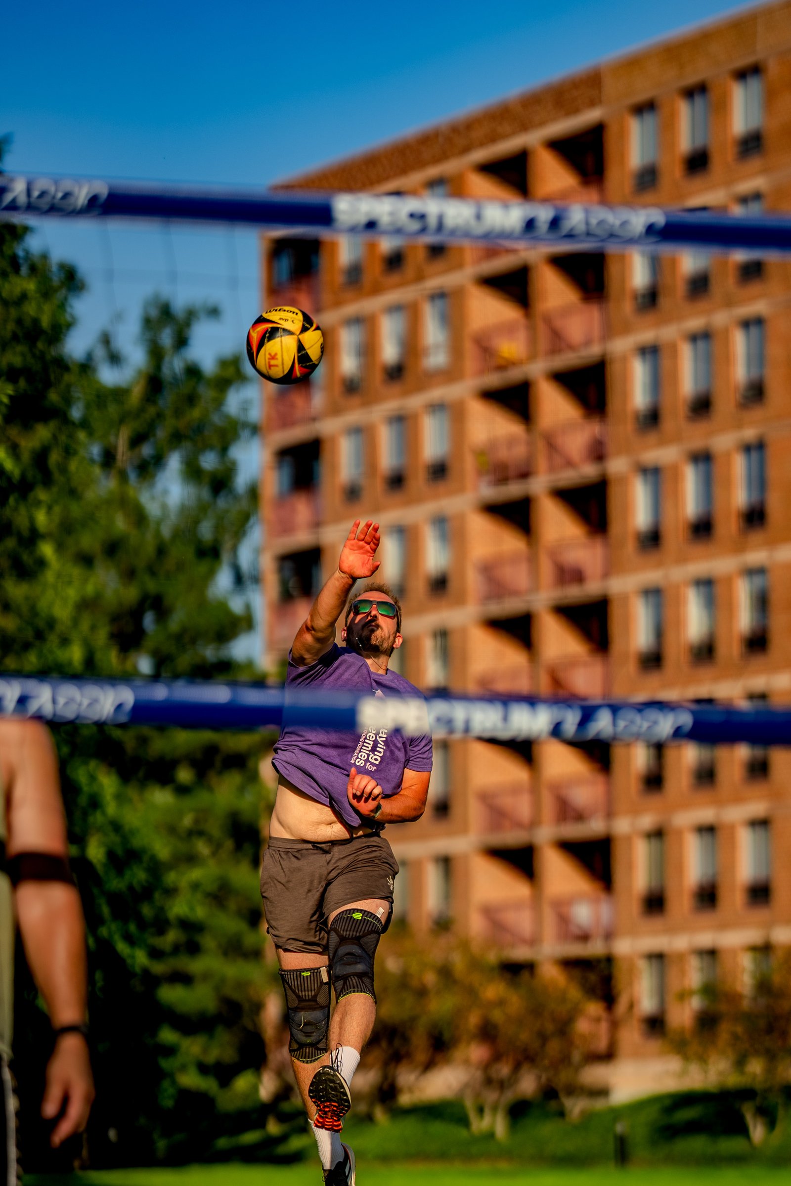 A player in a purple shirt and grey shorts jumps to hit a volleyball over the net, with a building in the background.