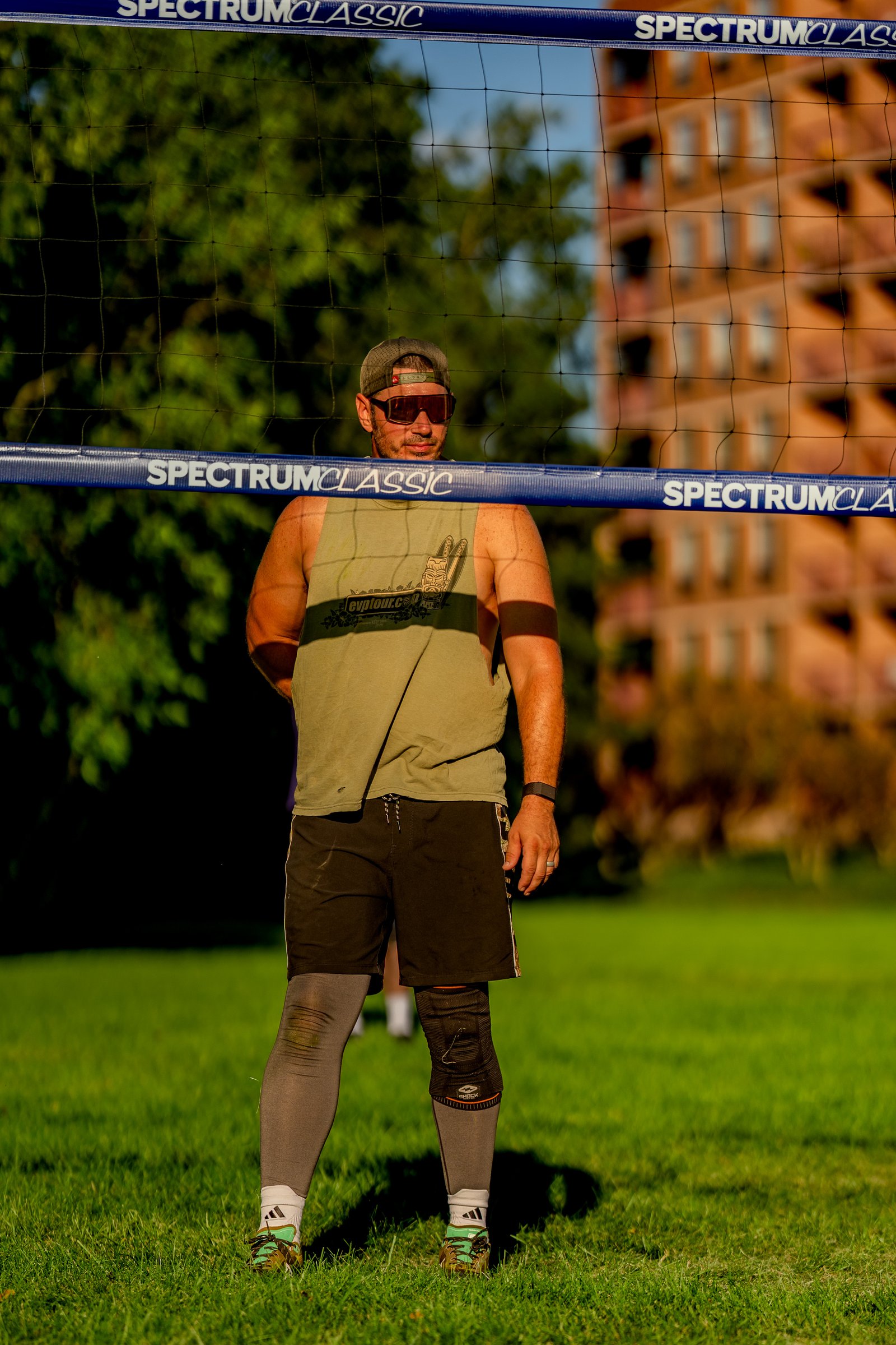 A player in a green tank top and black shorts stands on the grass near a volleyball net.