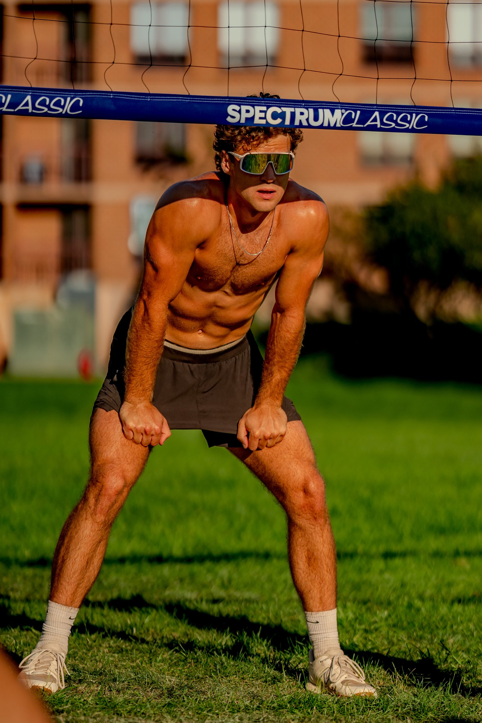 A player in dark shorts and white socks crouches on the grass, ready to play volleyball.