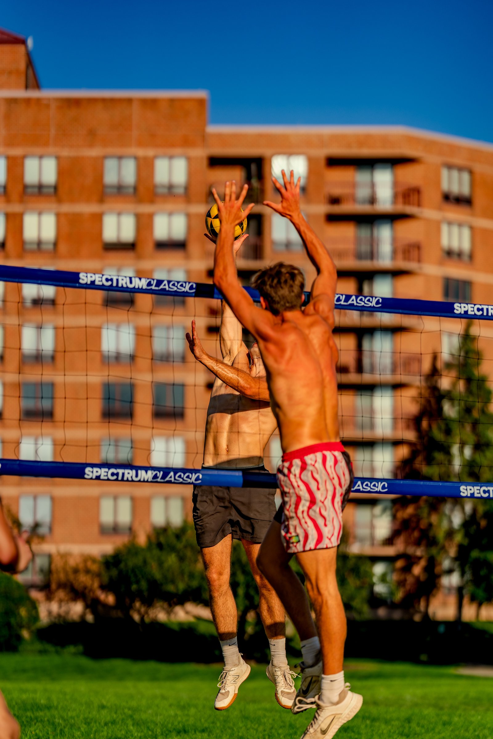 A player in red and white shorts jumps to hit a volleyball as another player in black shorts reaches up.