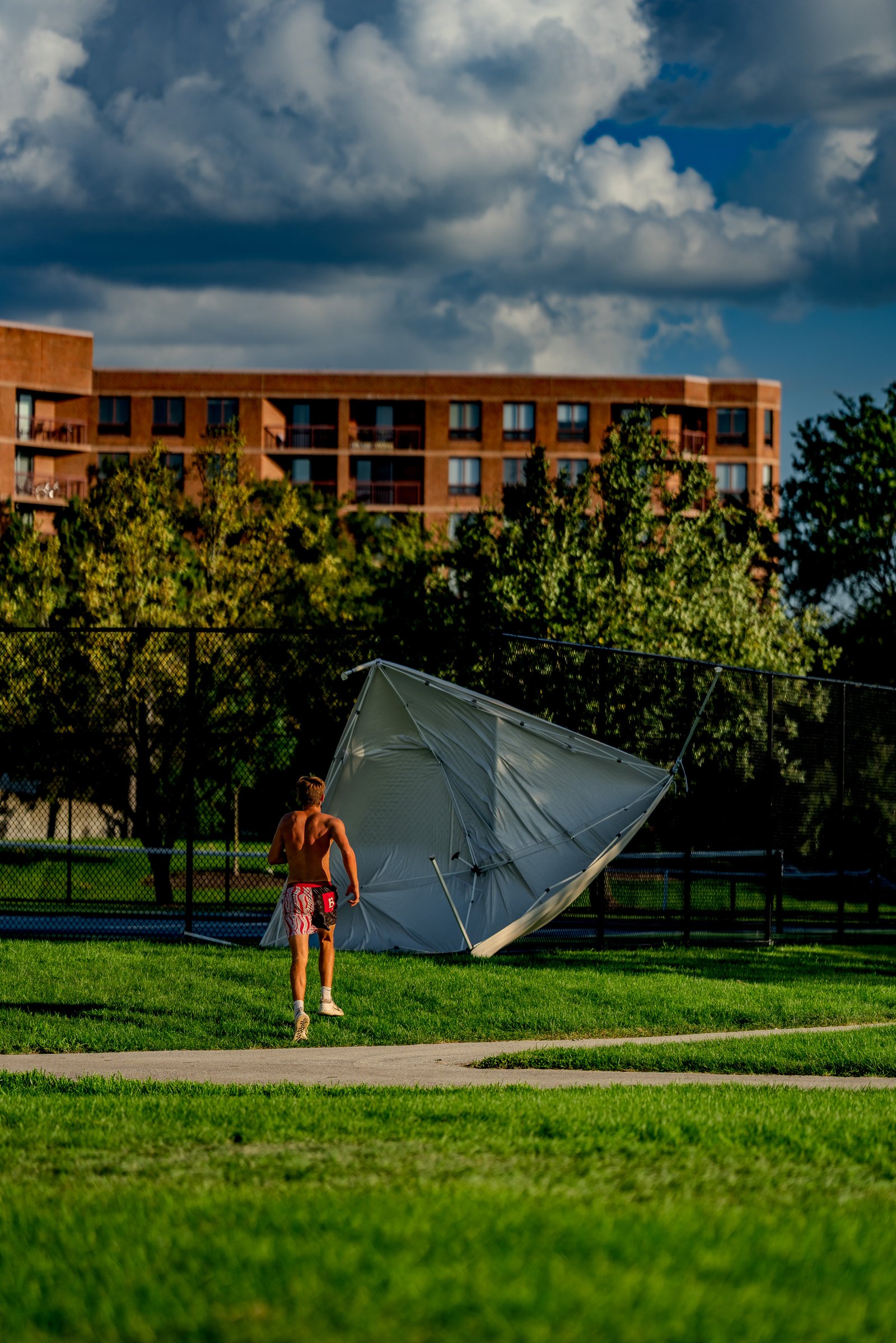 A player in red and black shorts runs past a collapsed tent on a grassy field with a building in the background.