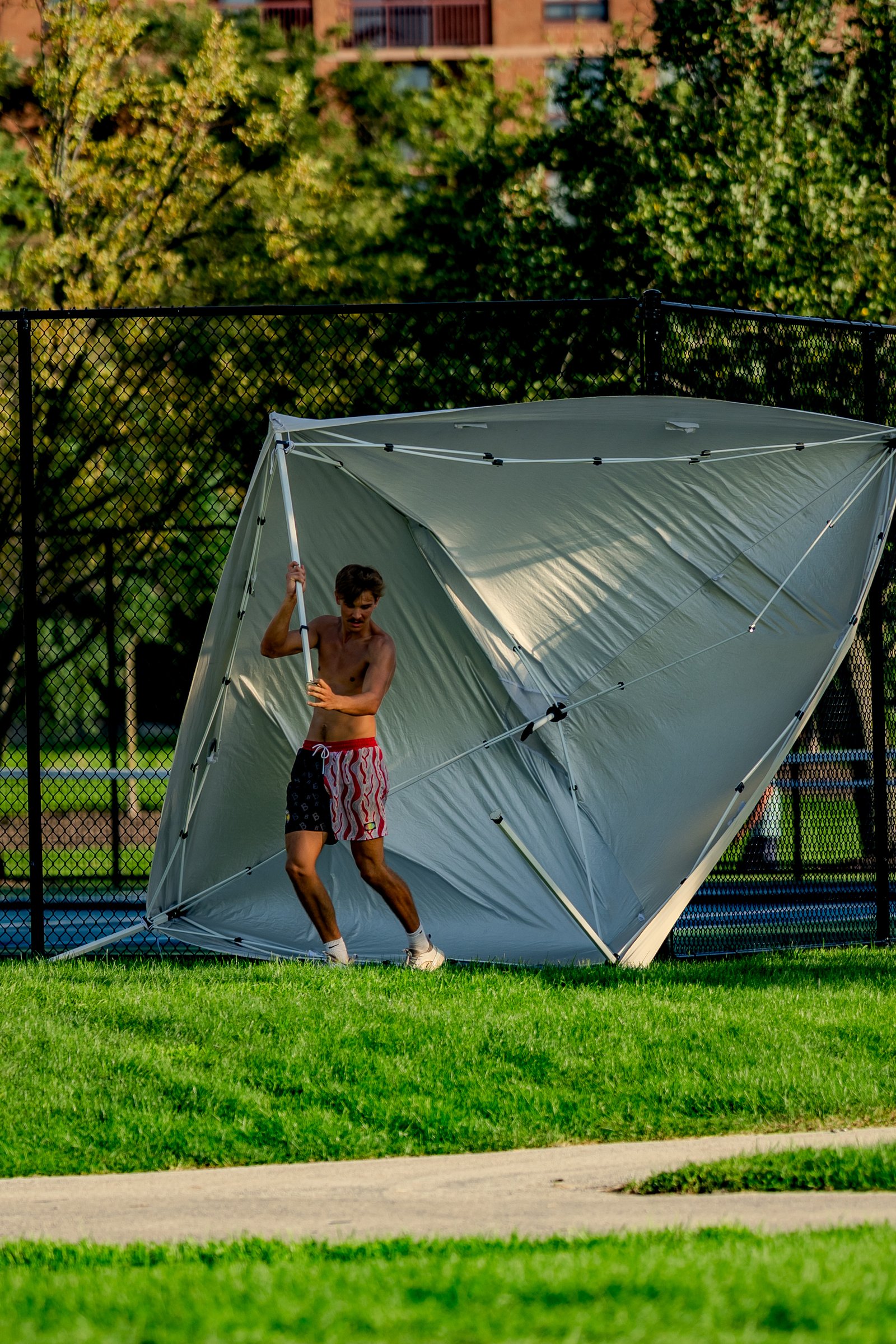 A player in patterned shorts struggles with a collapsed tent near a chain-link fence and trees.