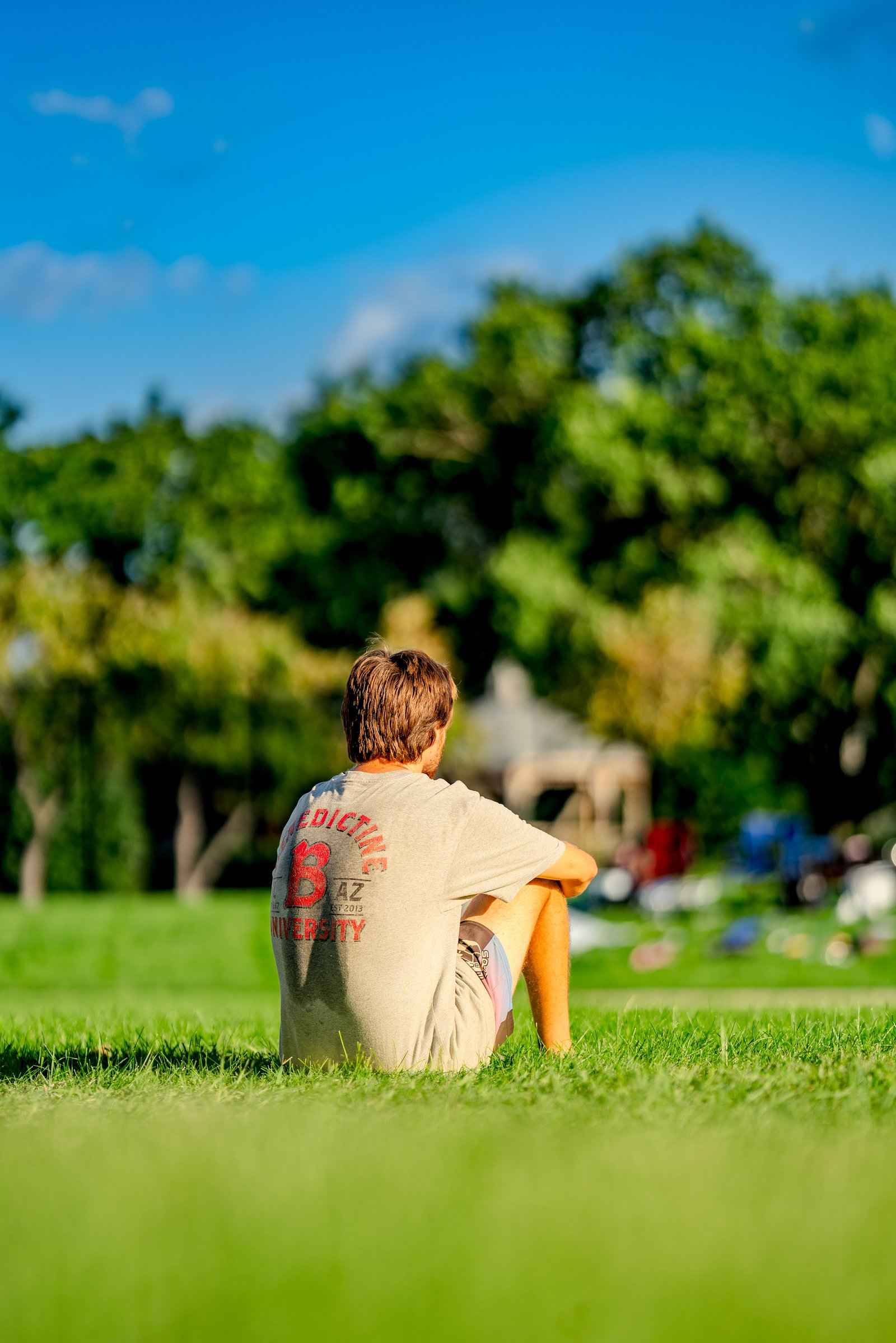 A player in a grey shirt sits on the grass at a tournament, with trees and a blue sky in the background.