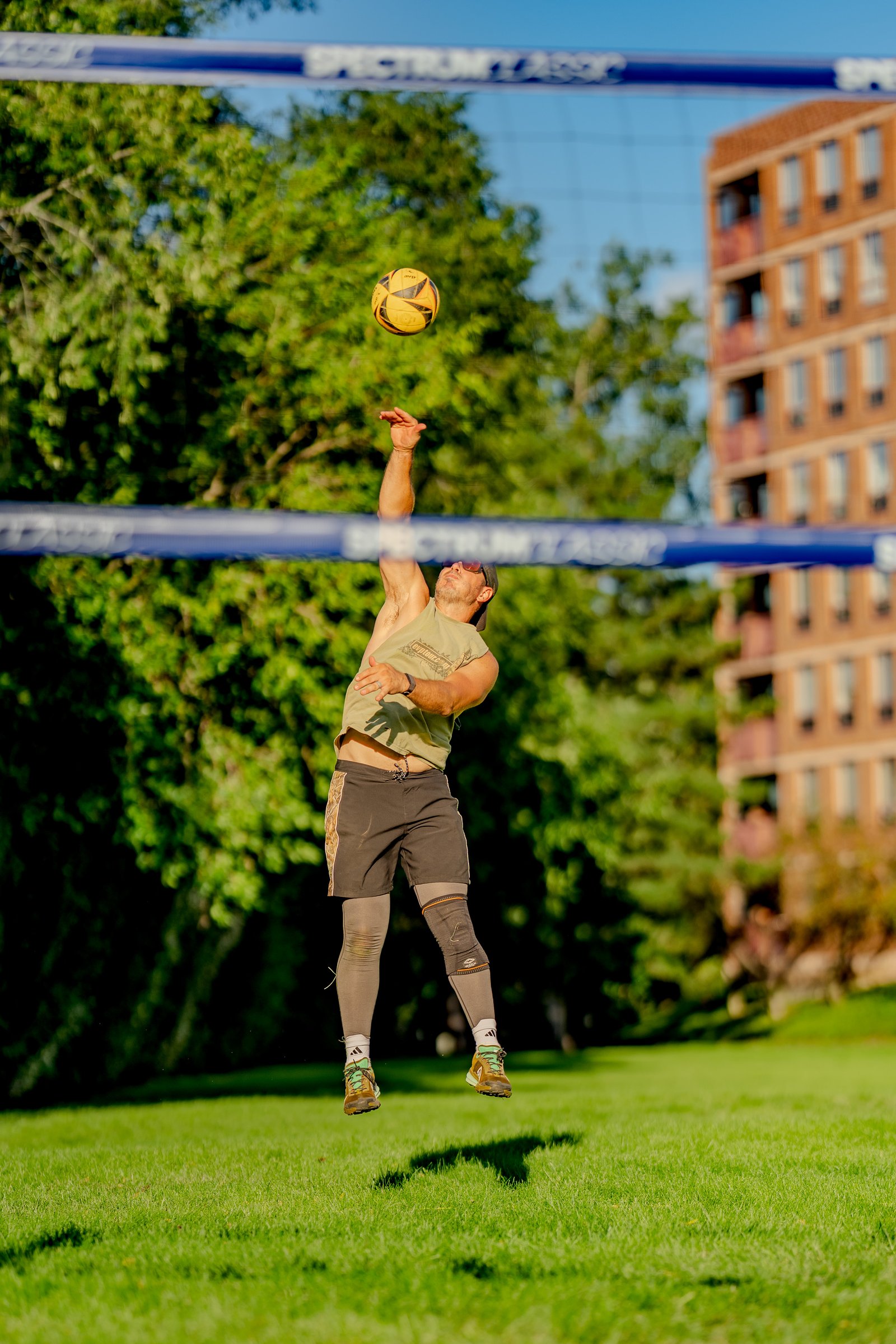 A player in a tan shirt and black shorts jumps to serve a volleyball over the net on a sunny day.