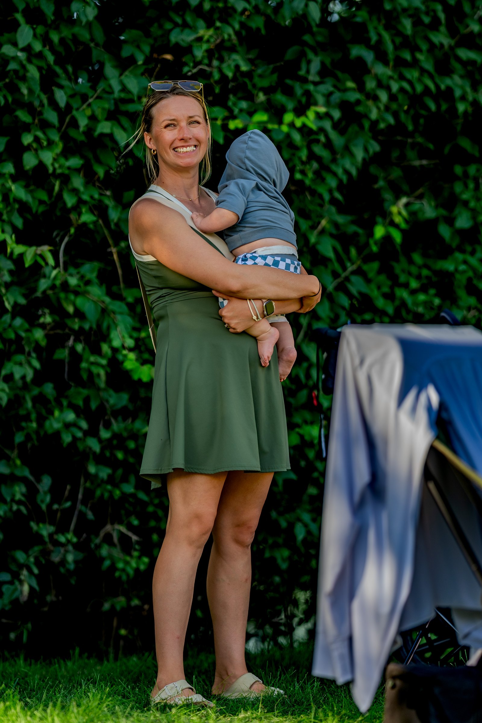 A smiling woman in a green dress holds a baby in a blue hoodie and checkered shorts.