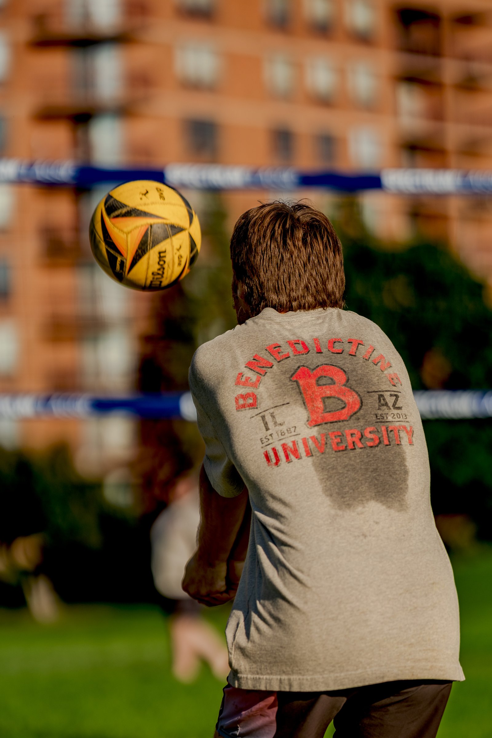 A player in a grey shirt with red lettering prepares to hit a yellow volleyball during a tournament.