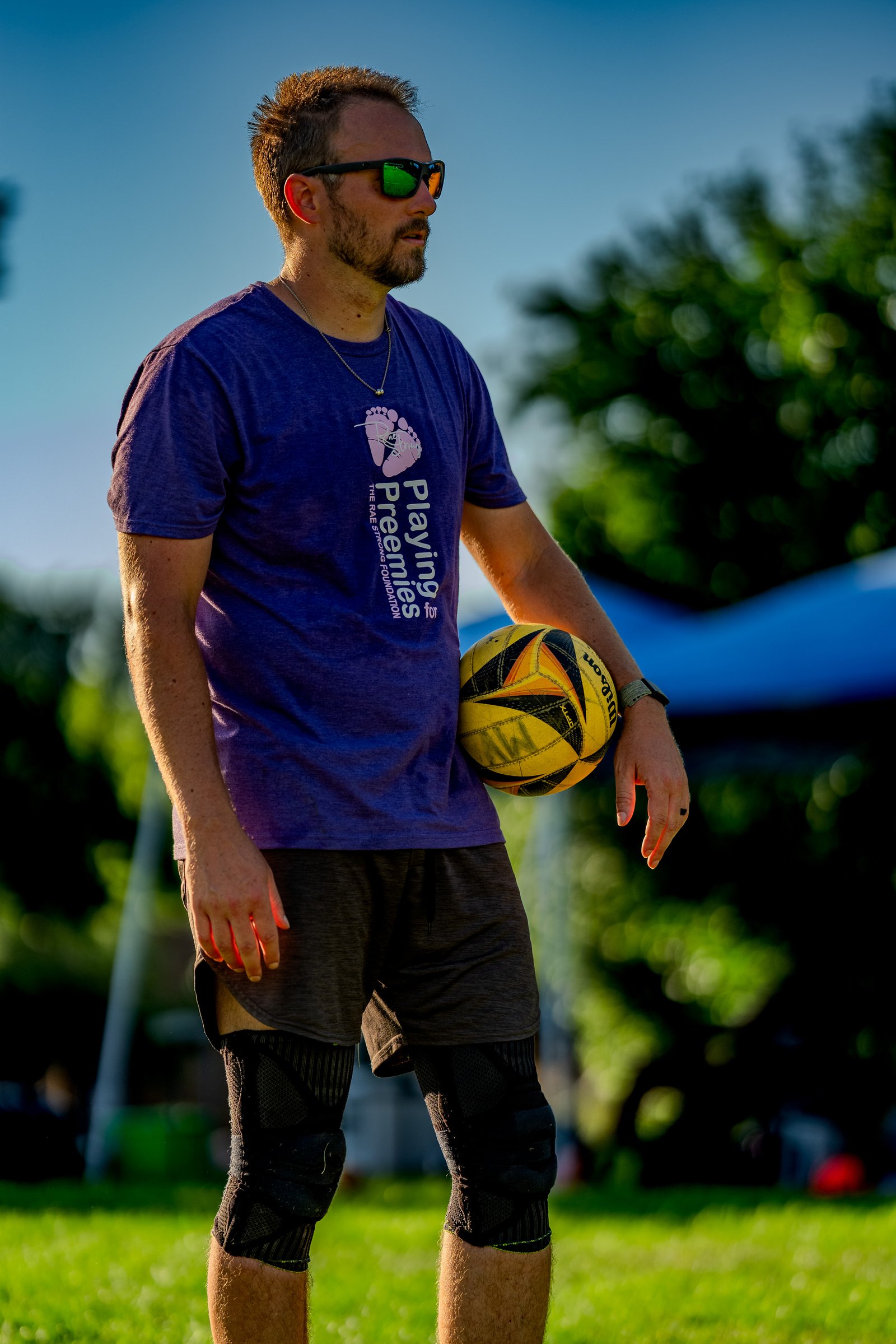 A player in a purple shirt and dark shorts holds a volleyball, wearing knee pads and sunglasses.