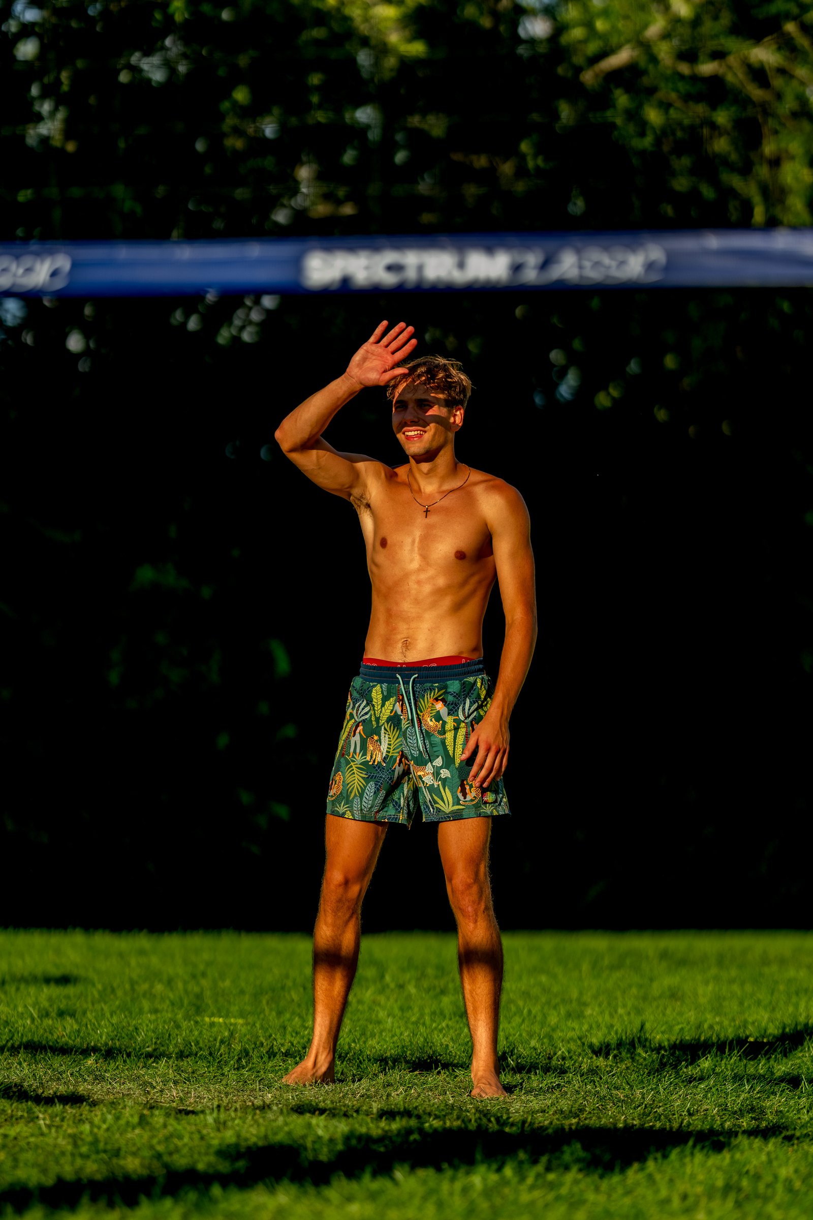 A player in patterned shorts shields his eyes from the sun on a grassy court near a volleyball net.