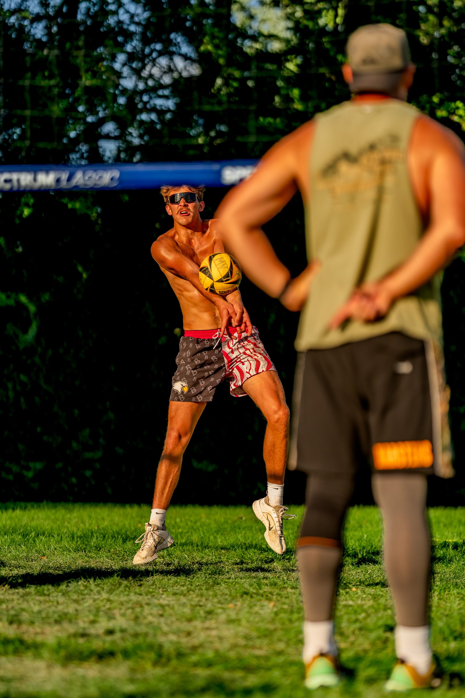 A player in patterned shorts passes a volleyball during a tournament, with another player in a green top facing away.