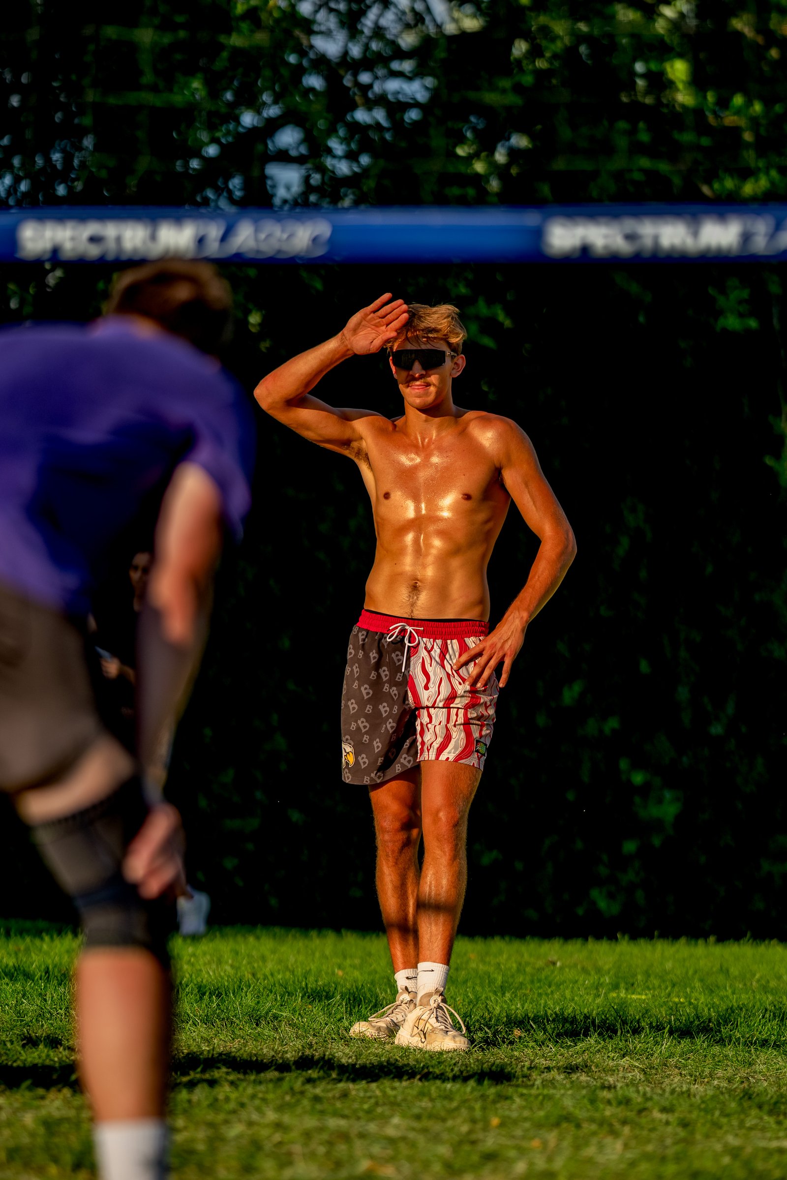 A player in red and white patterned shorts shields his eyes from the sun while another player in a purple shirt bends down on the grass court.