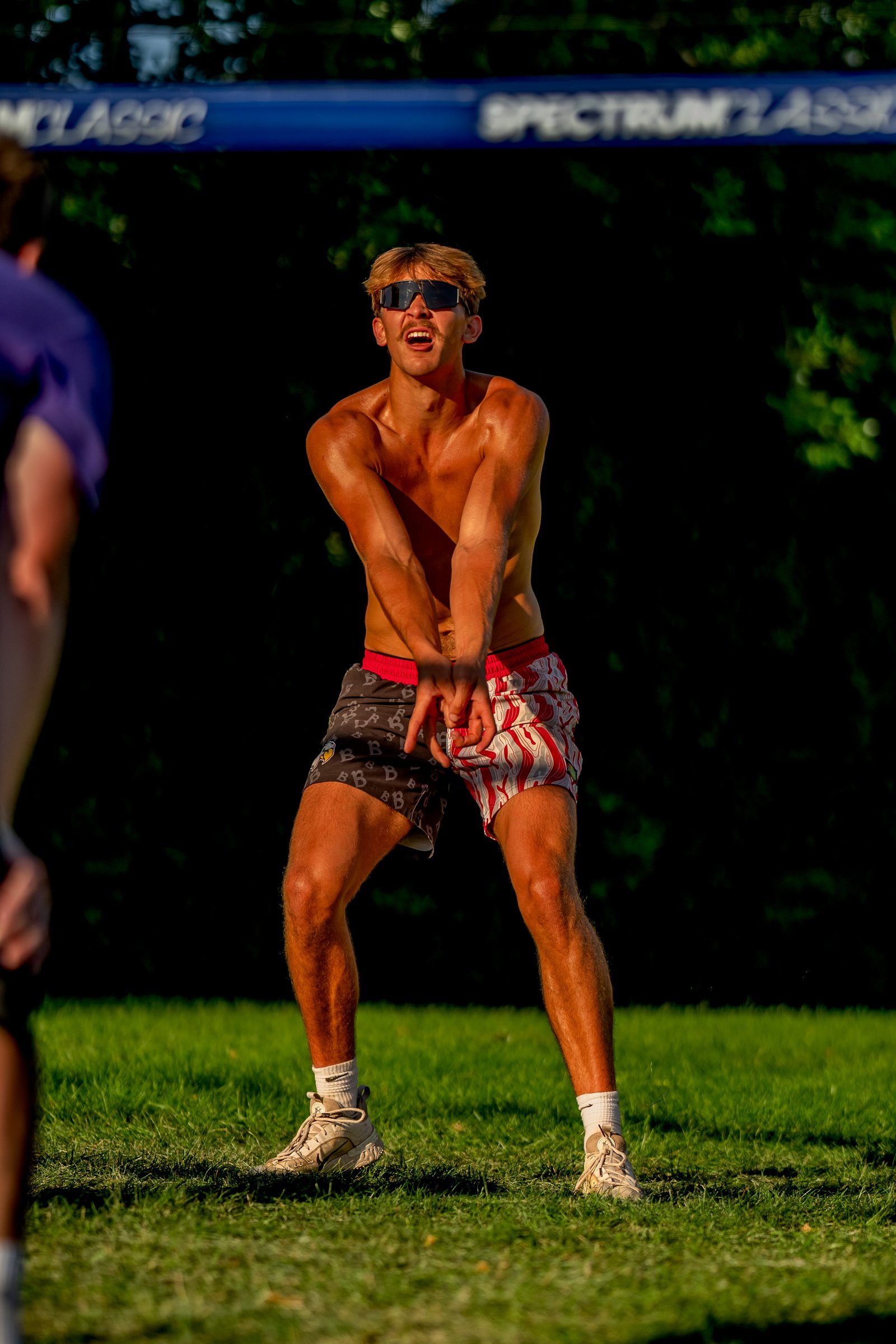 A player in patterned shorts prepares to pass a volleyball on a grassy court with a blue net overhead.