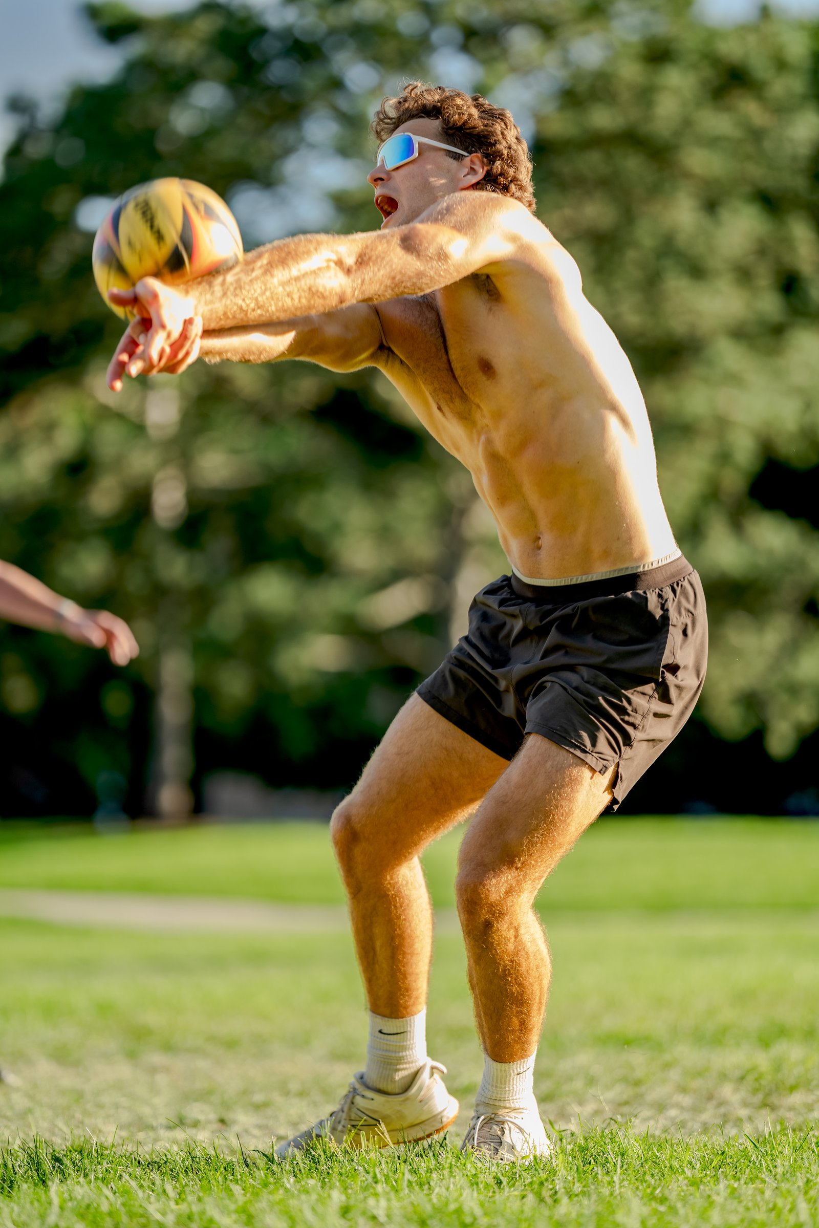 A player in black shorts and white sunglasses passes a volleyball on a grassy field.