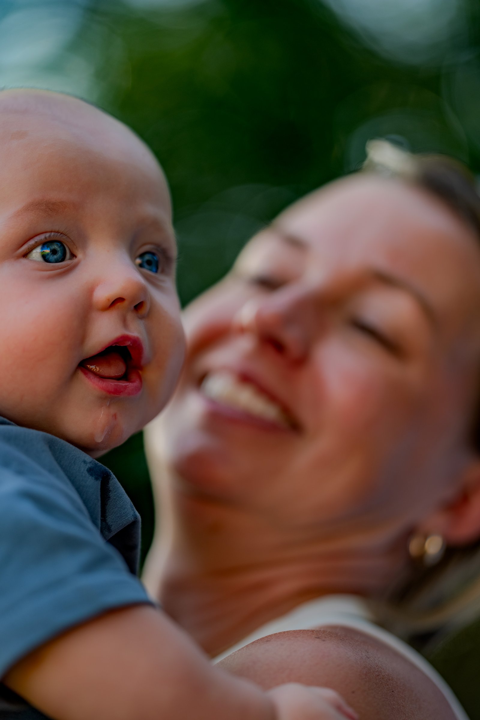 A baby with blue eyes looks to the side while being held by a smiling person in a white top.