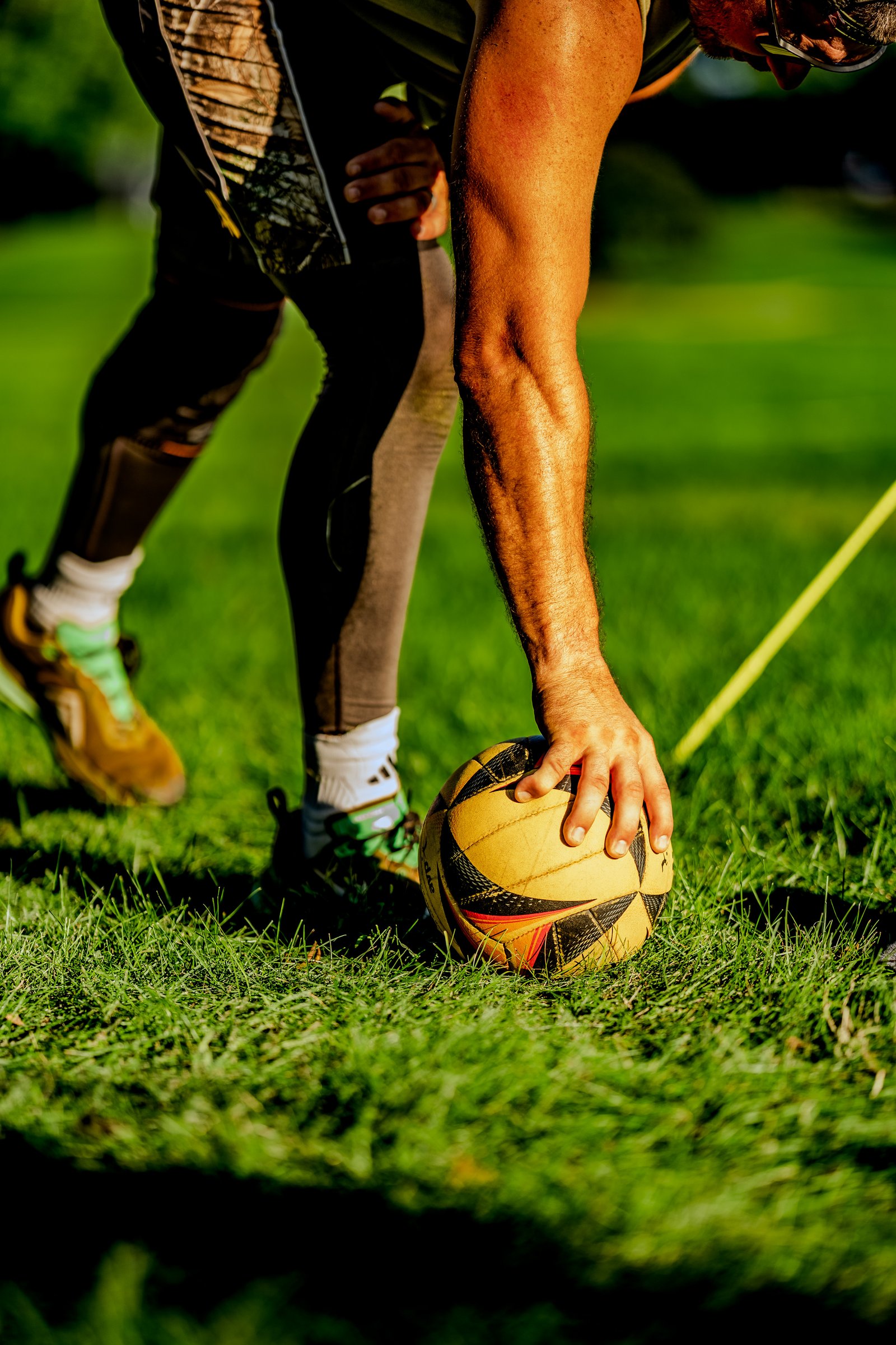A player in a black top and camouflage shorts reaches for a volleyball on the grass.