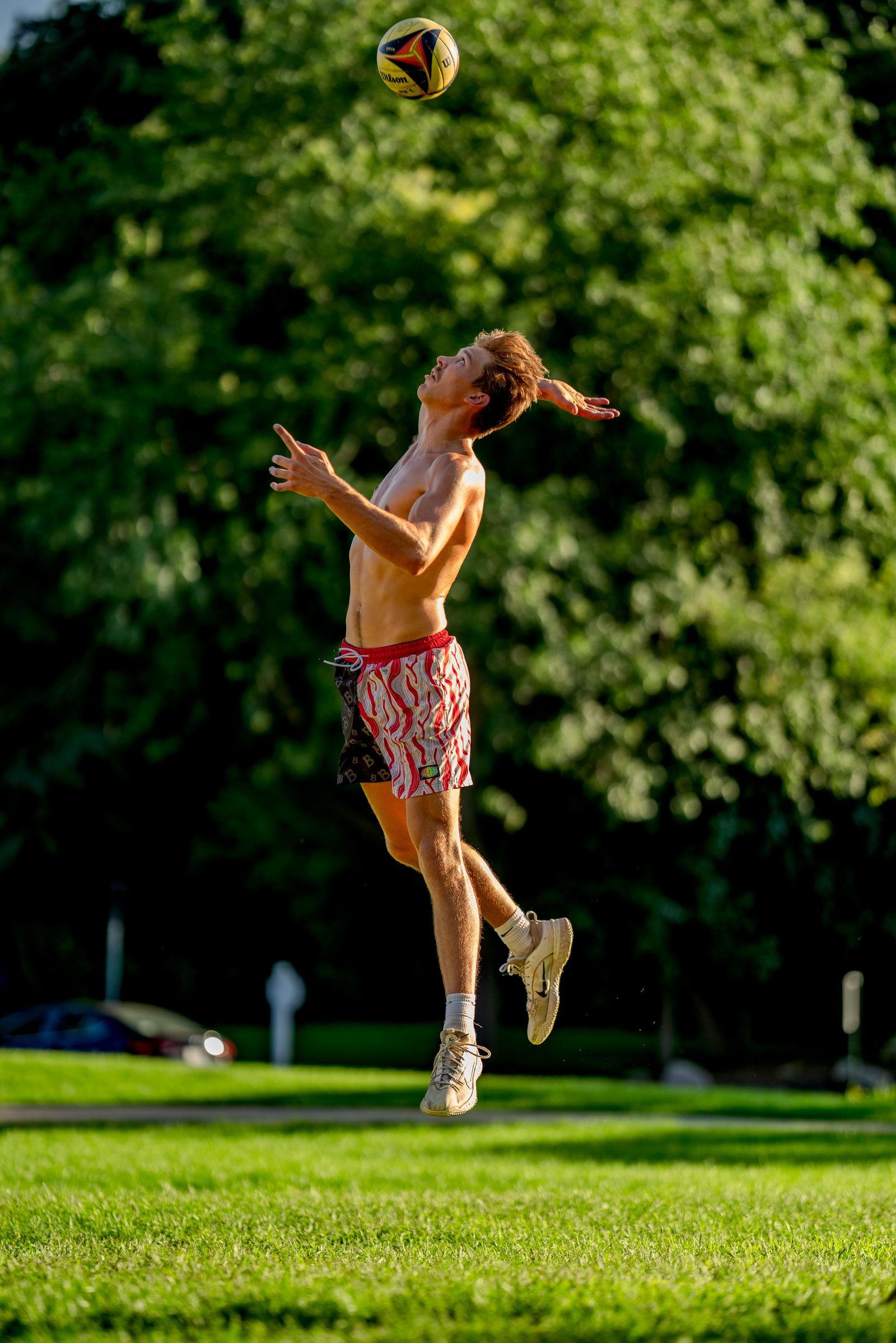 A player in red and white patterned shorts jumps to hit a volleyball outdoors with trees in the background.