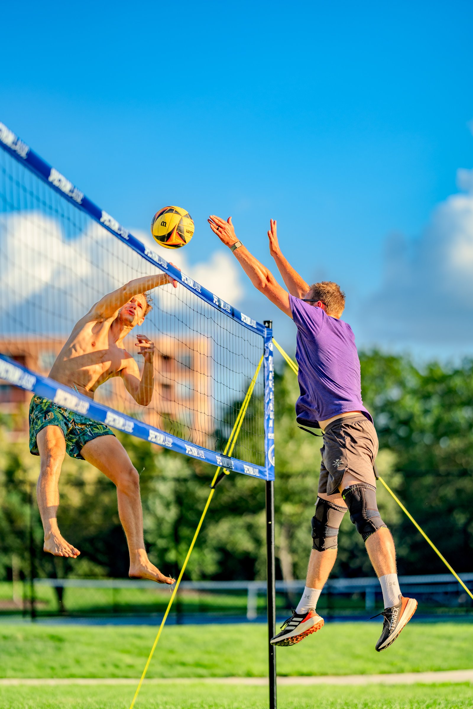 A player in green shorts jumps to hit the ball over the net while a player in a purple shirt reaches up to block during a volleyball match.