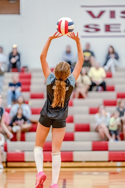 A player in a black top and blue sleeves sets a volleyball in a gymnasium with spectators in the background.