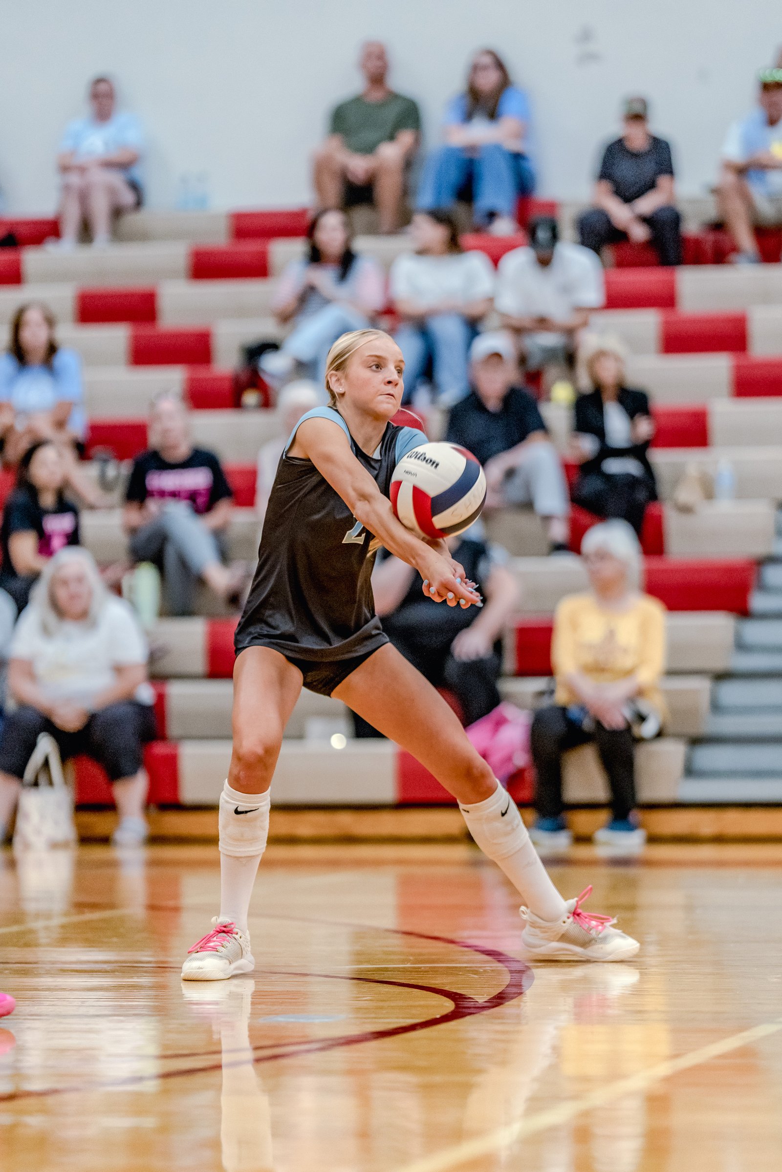 A player in a black and light blue jersey passes a volleyball on the court with spectators in the background.
