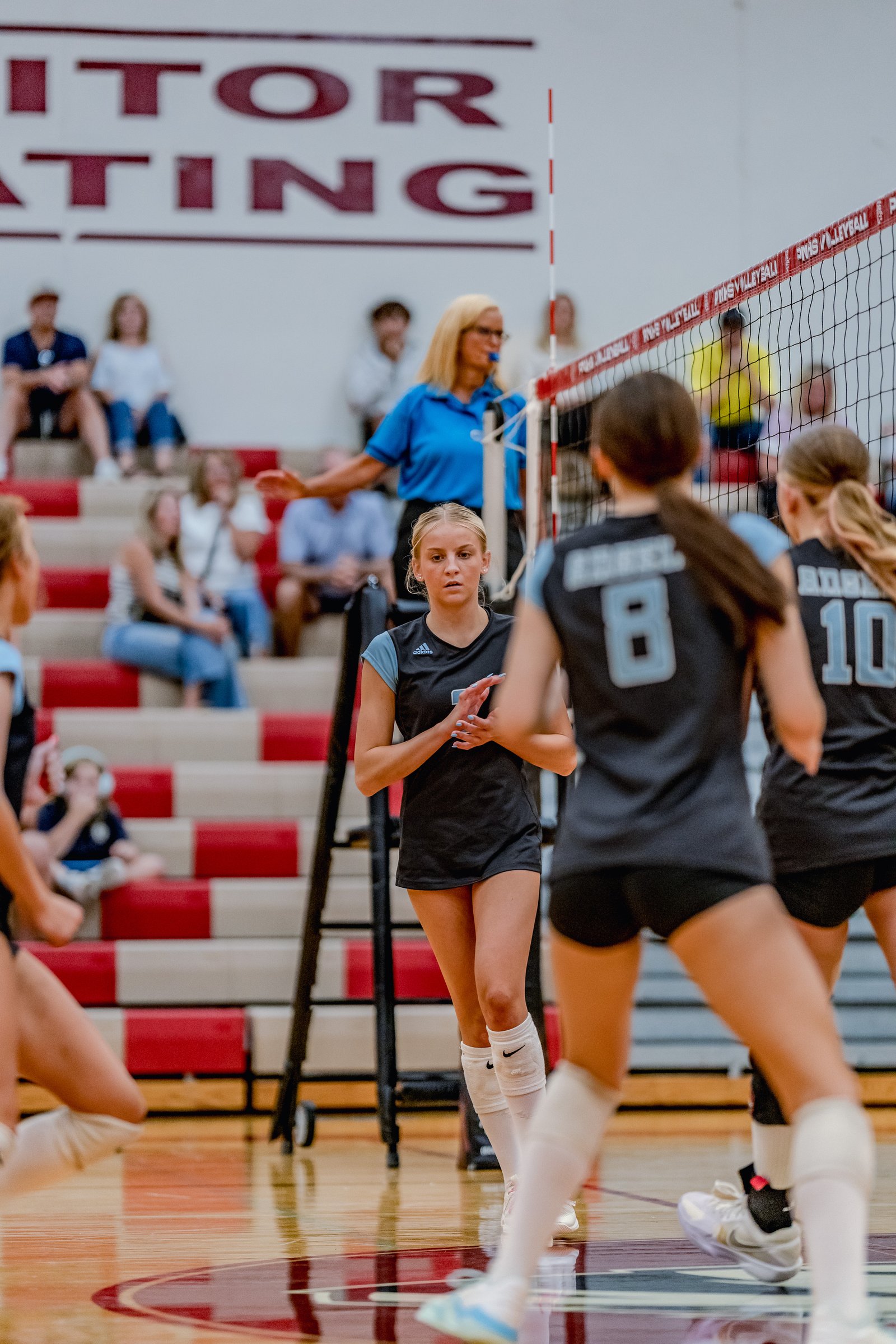 A player in a black top with light blue sleeves prepares to play volleyball, with teammates in black and light blue jerseys visible.