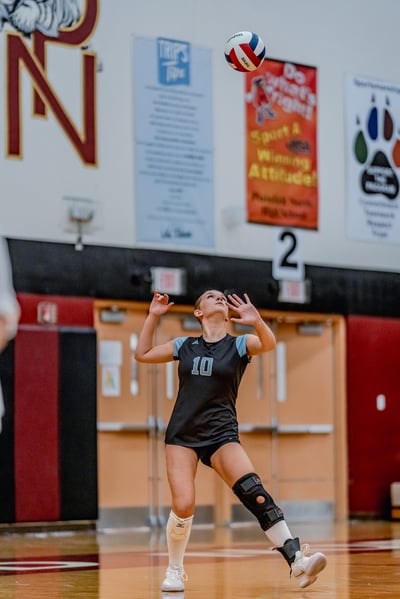A player in a black jersey with light blue accents and number 10 prepares to hit a volleyball in a gymnasium.