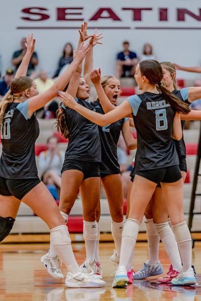 Players in black and light blue jerseys celebrate with hands raised on the court, with one player in jersey number 2 shouting.