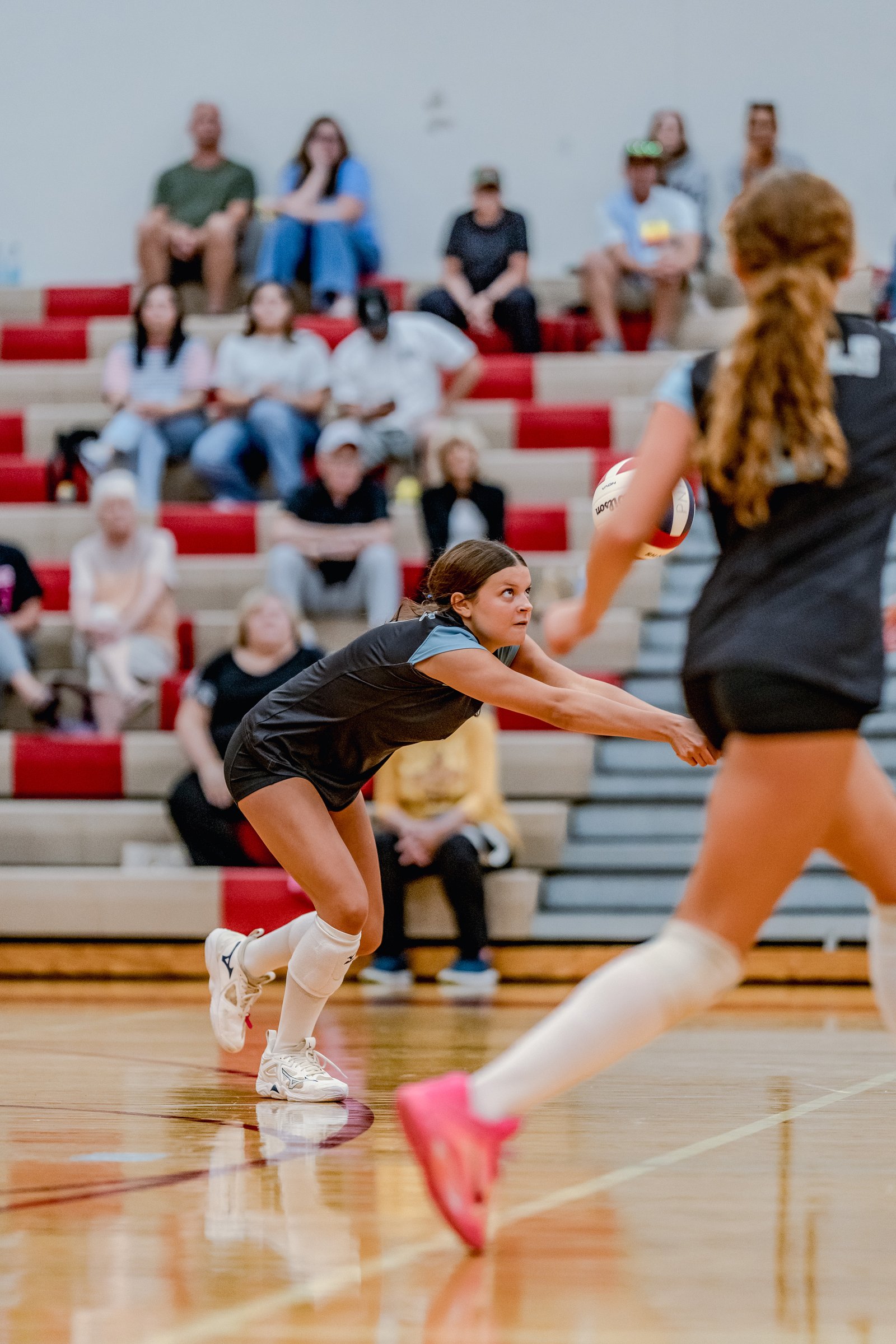 A player in a black top and shorts passes a volleyball on the court with spectators in the background.