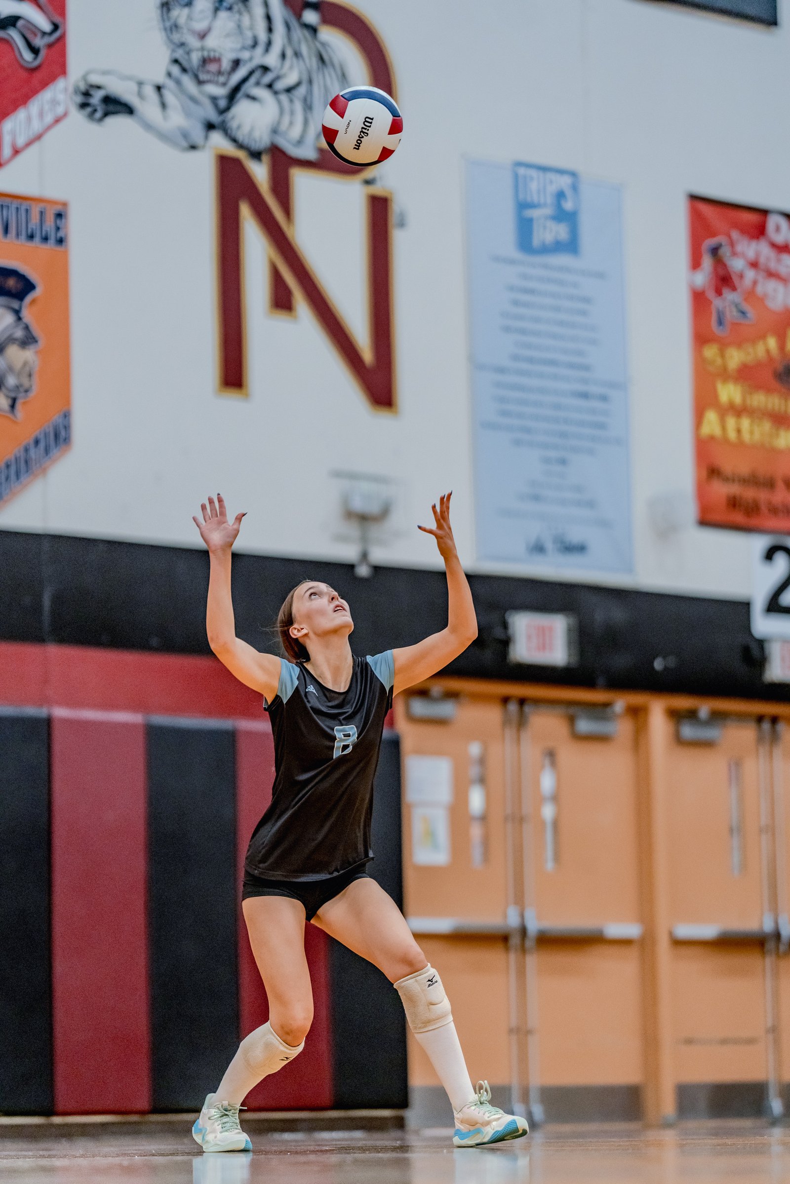 A player in a black top with light blue accents prepares to set a volleyball in a gymnasium.