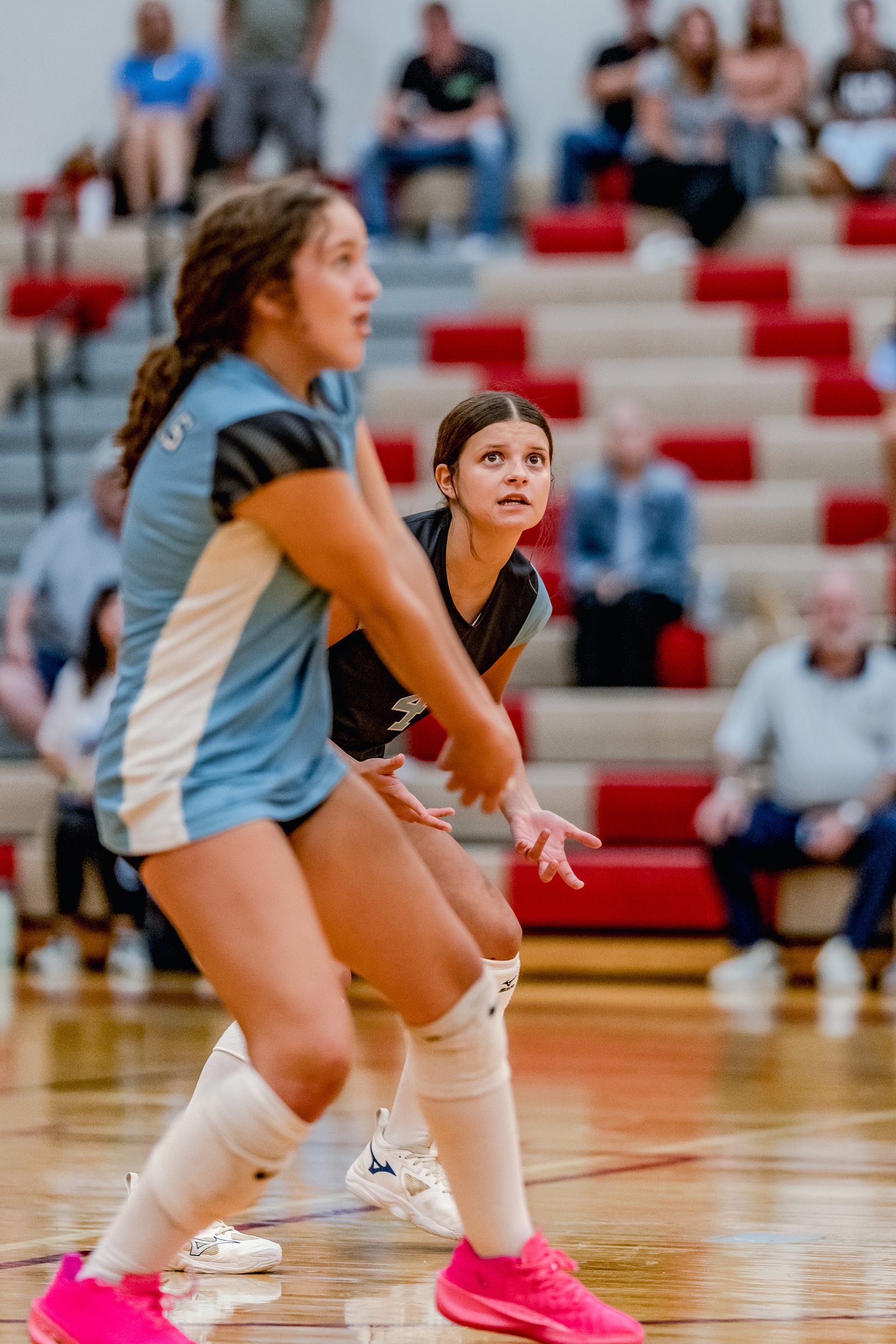 A player in a light blue jersey with white trim and a player in a dark jersey with white trim prepare to play volleyball on a court.