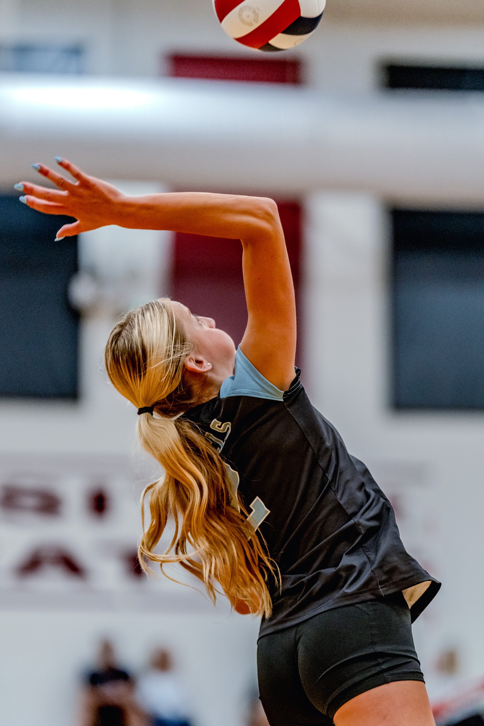 A player in a black jersey with light blue trim serves a volleyball during a game.