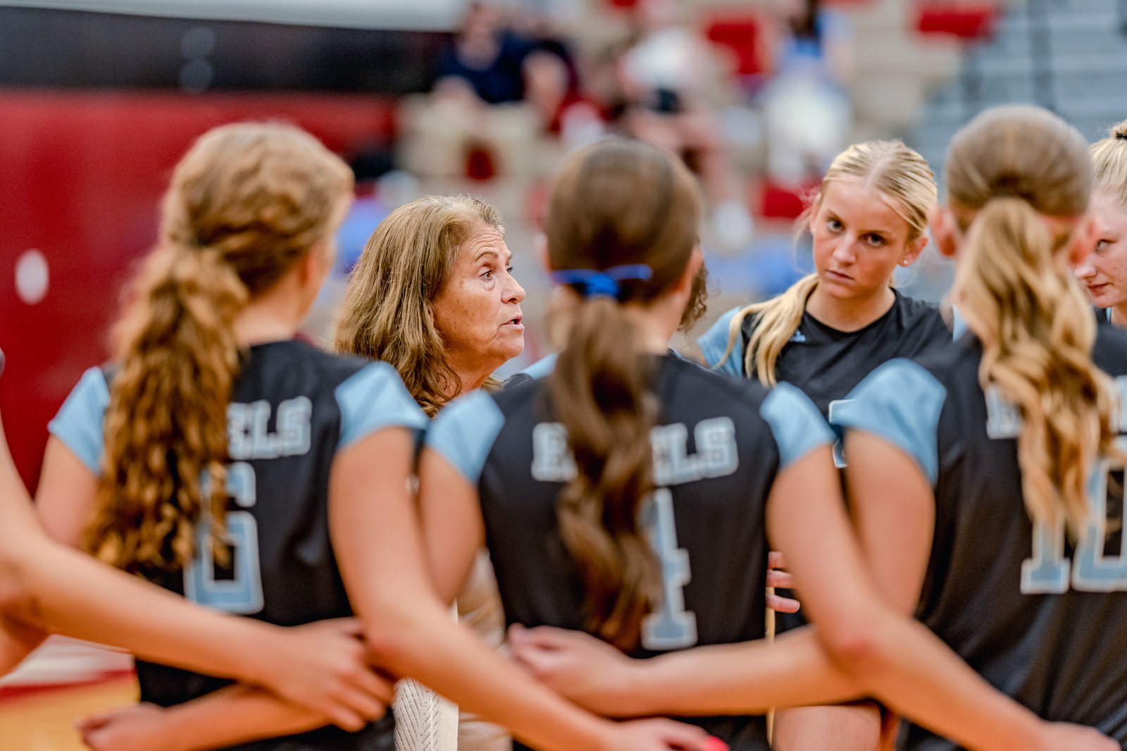 A coach speaks to players in black and light blue jerseys during a huddle on the court.