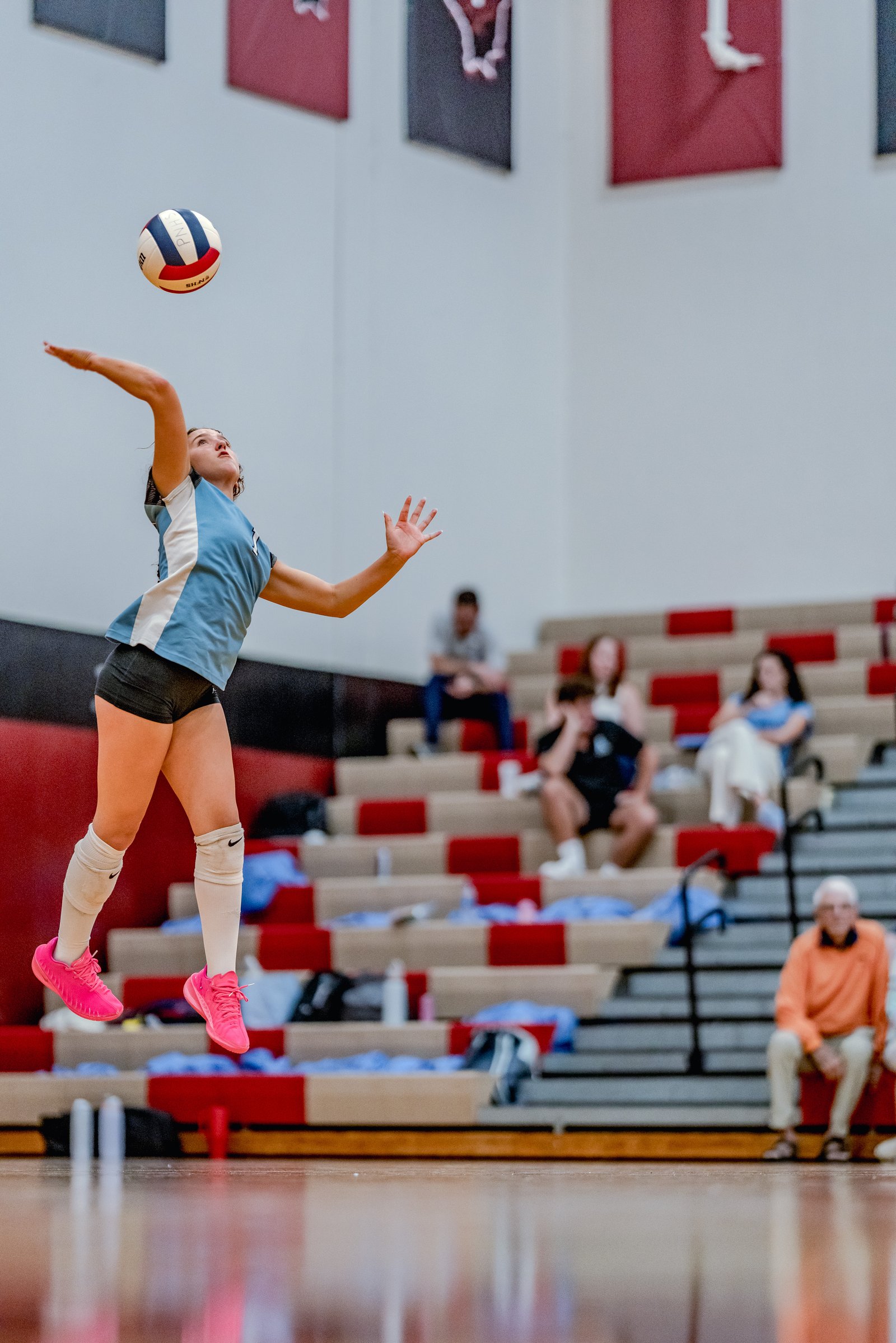 A player in a light blue jersey serves a volleyball during a game, with spectators in the background.