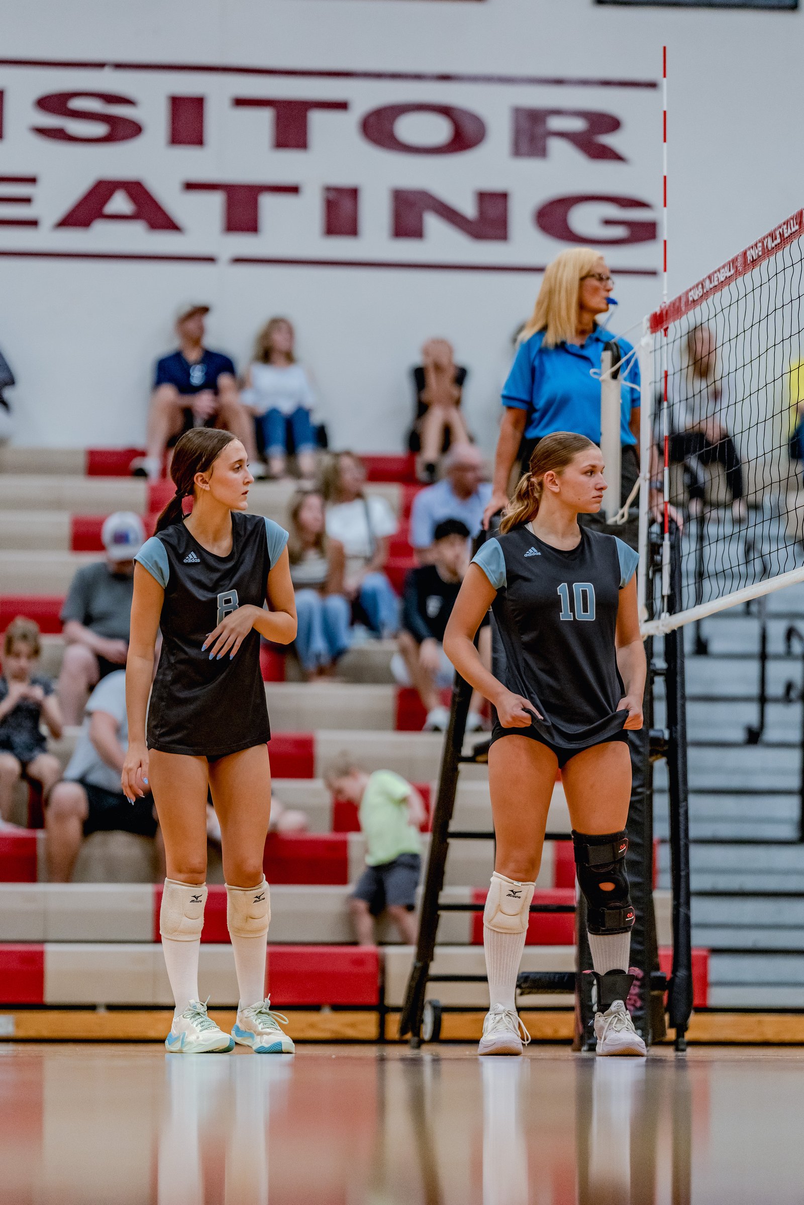 Two volleyball players in black and light blue uniforms stand on the court, with spectators in the background.