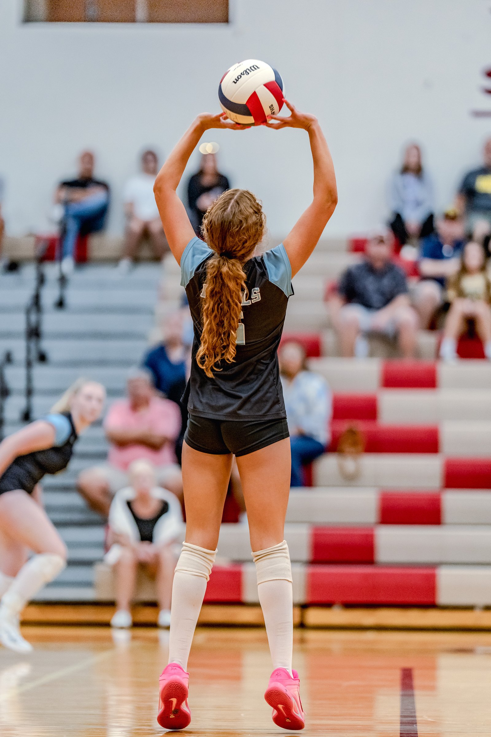 A player in a black and light blue jersey prepares to serve a volleyball in a gymnasium with spectators in the background.