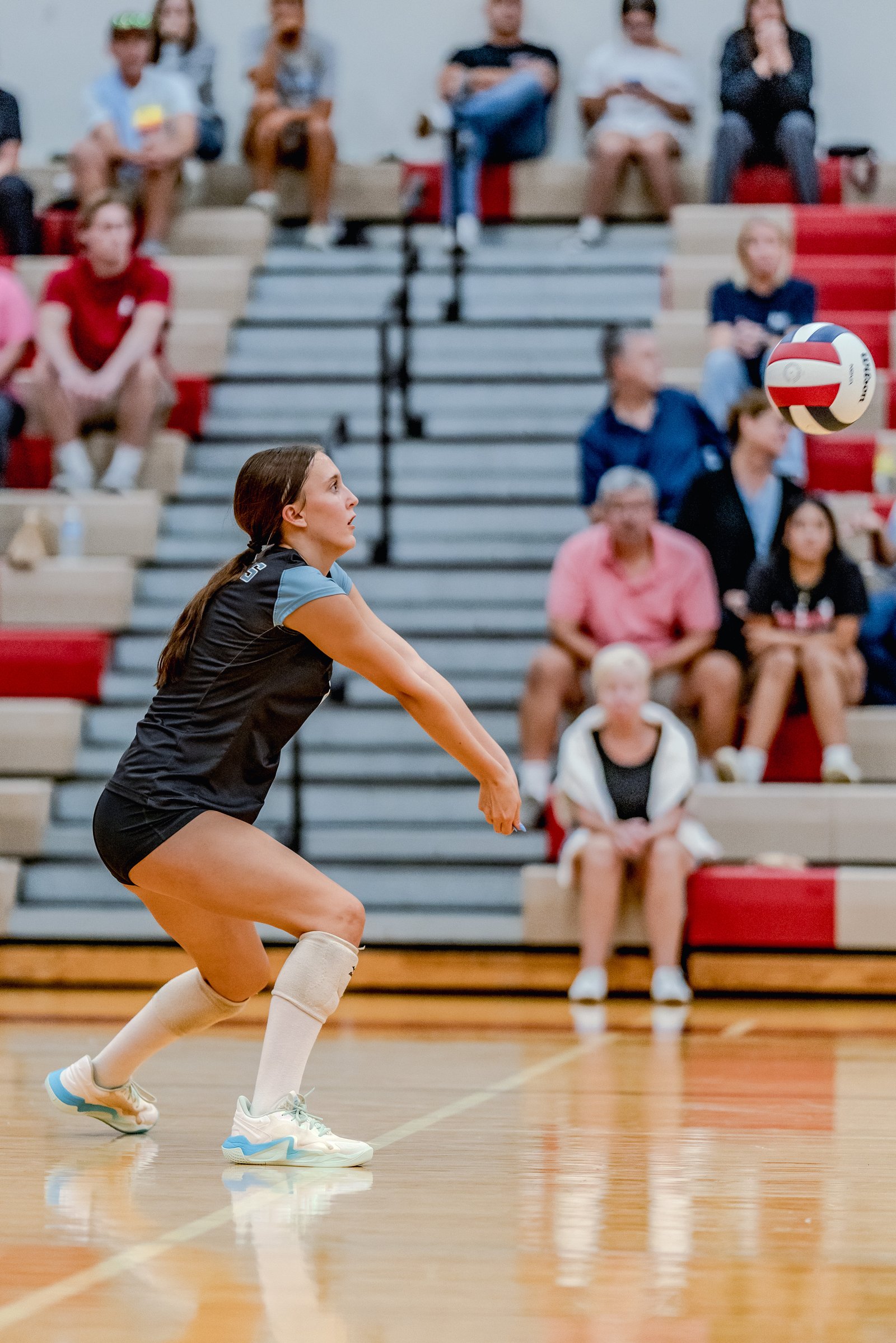A player in a black and light blue top prepares to pass a volleyball during a game with spectators in the background.