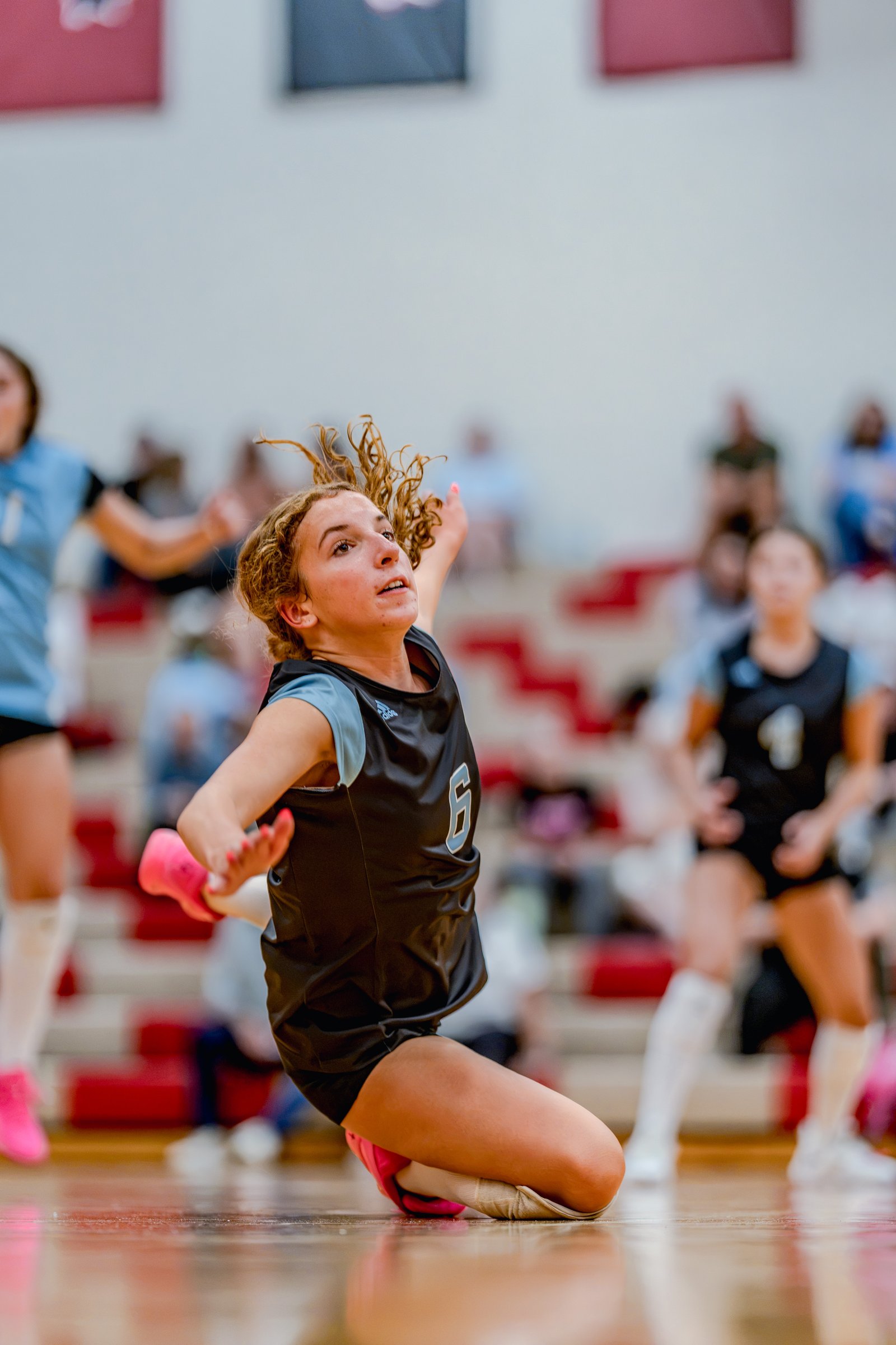 A player in a black and light blue jersey with number 6 dives for a volleyball on the court, with other players in the background.