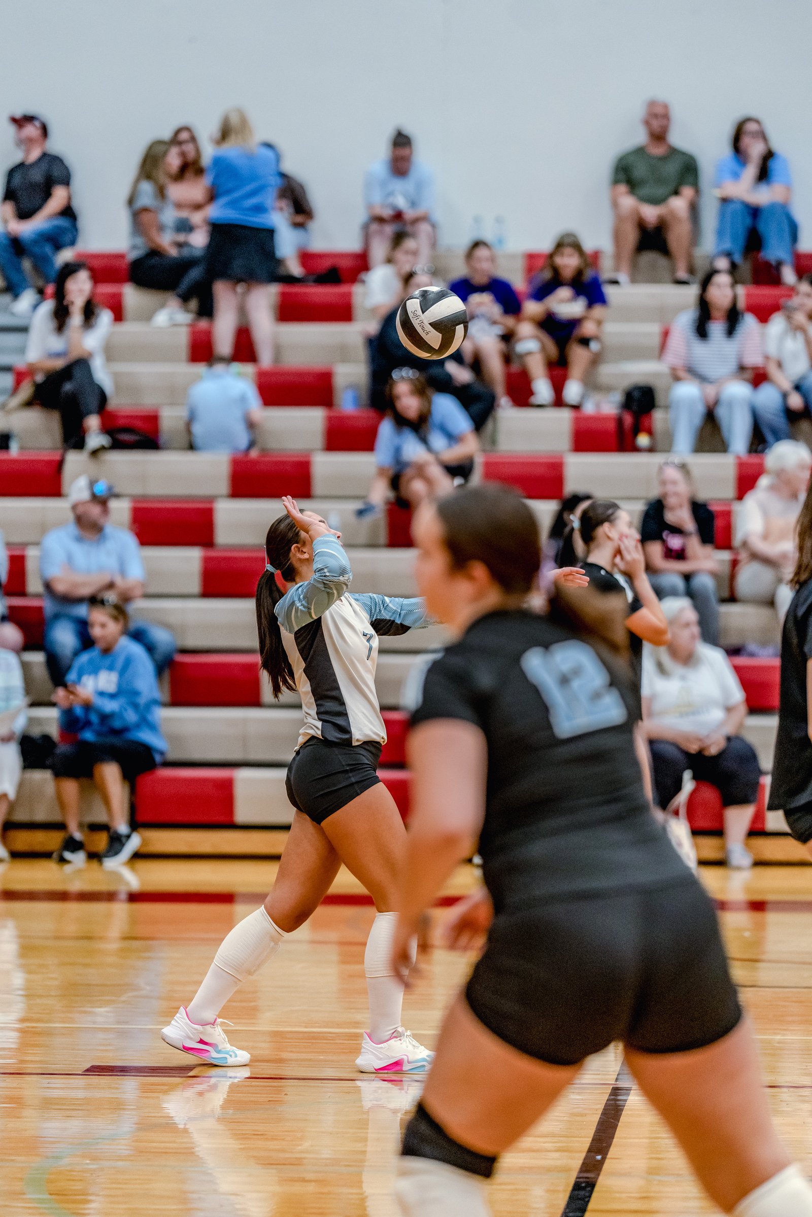 A player in a white and blue jersey serves a volleyball during a game with spectators in the background.