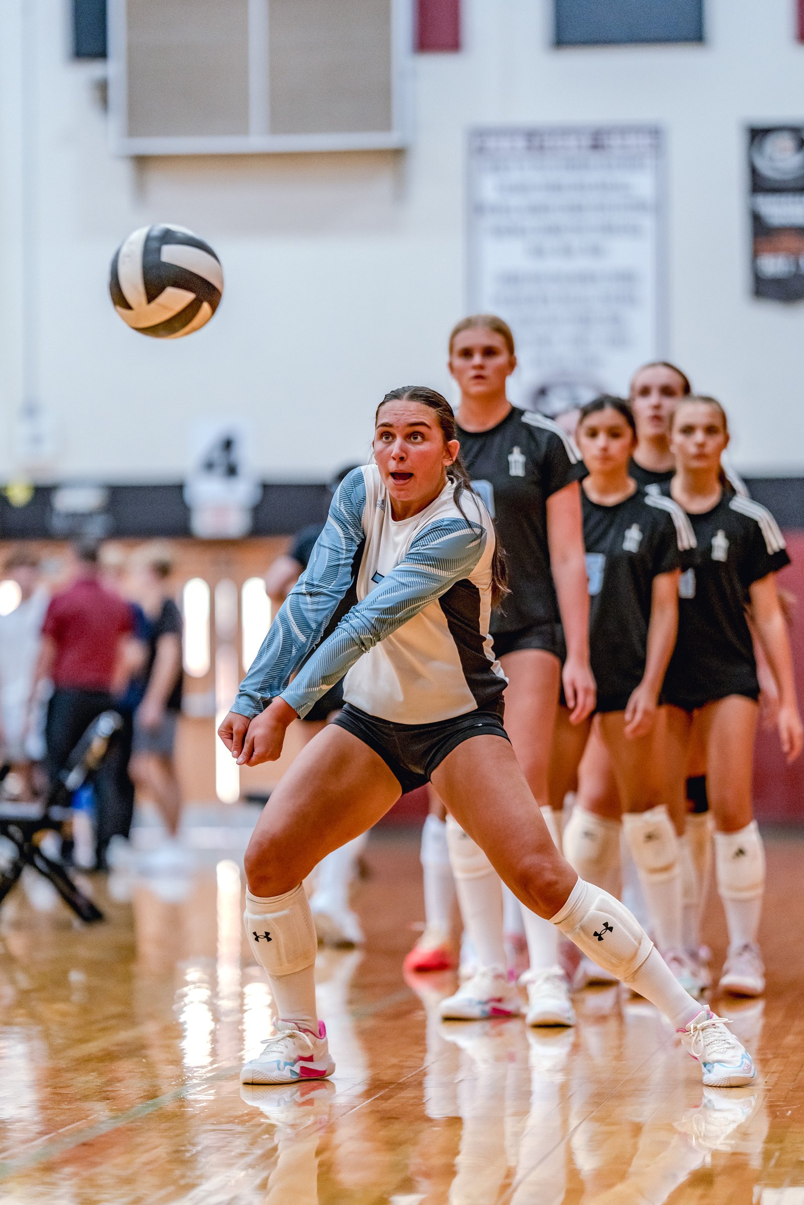 A player in a blue and white top prepares to pass a volleyball as teammates in black tops line up behind her on the court.
