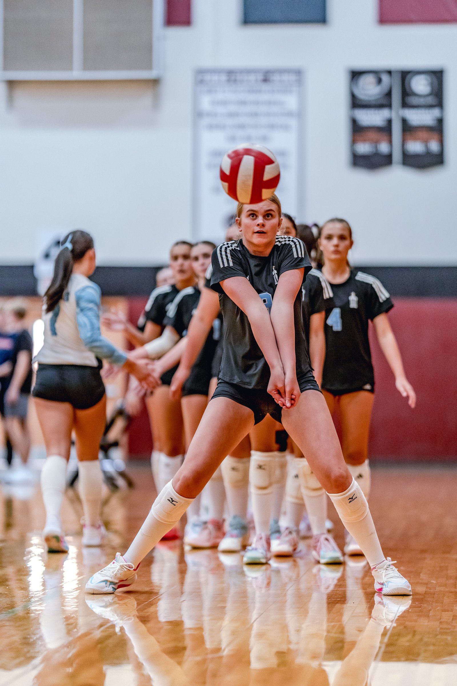 A player in black prepares to receive a volleyball, with teammates in black and a player in light blue behind her.
