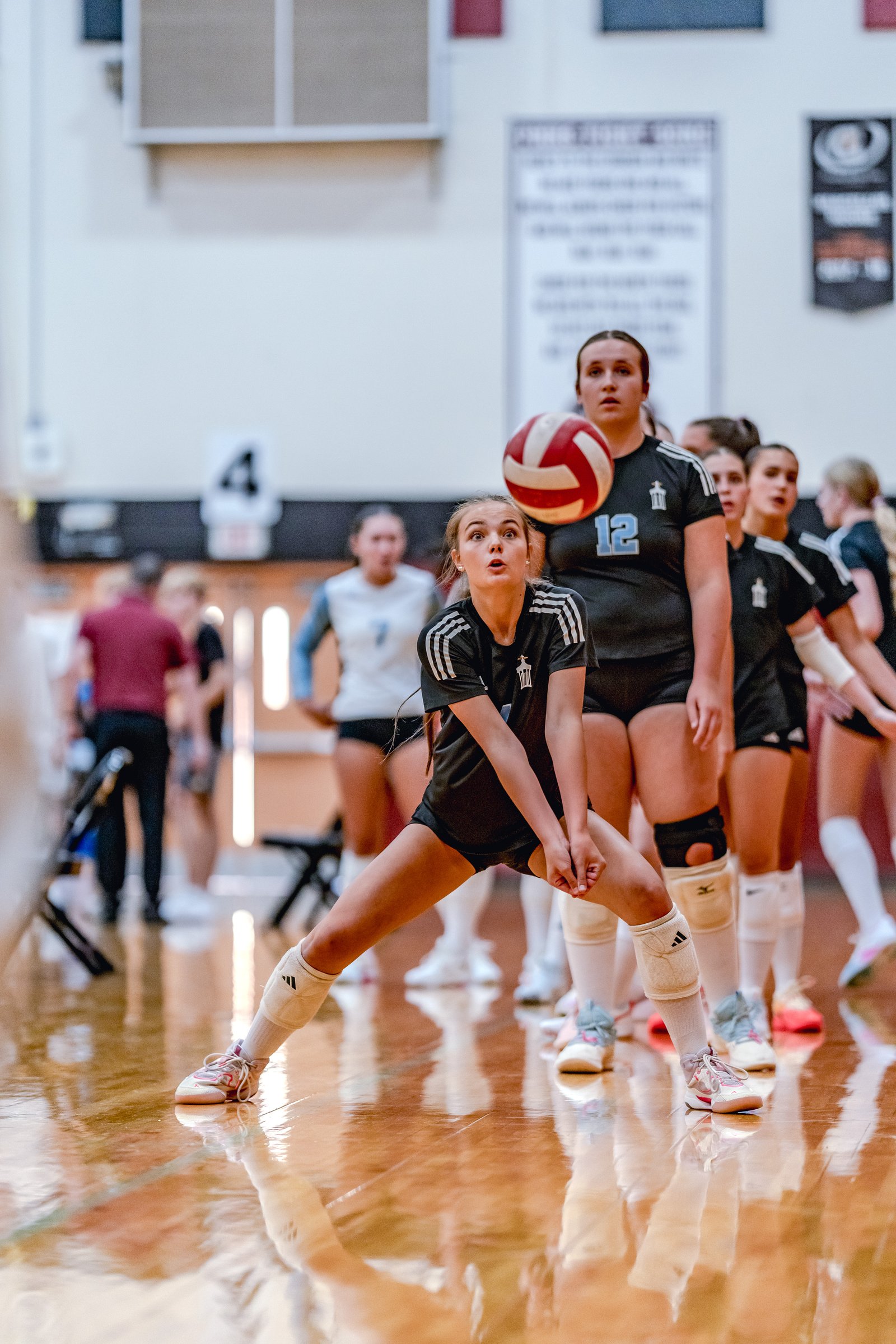 A player in black prepares to dig a volleyball as teammates in black and white look on during a game.