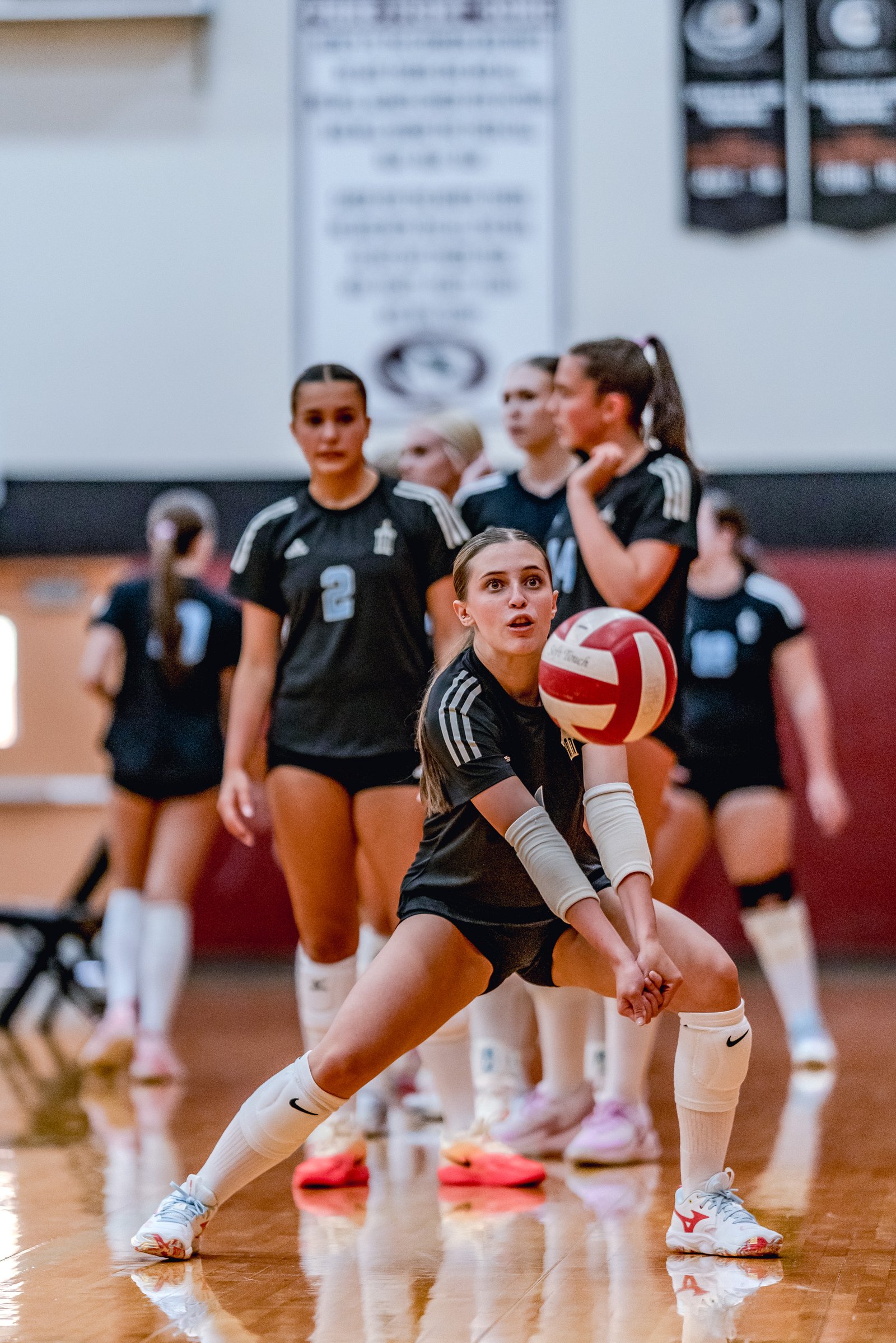 A player in a black top and white socks passes a volleyball while teammates in black uniforms watch.