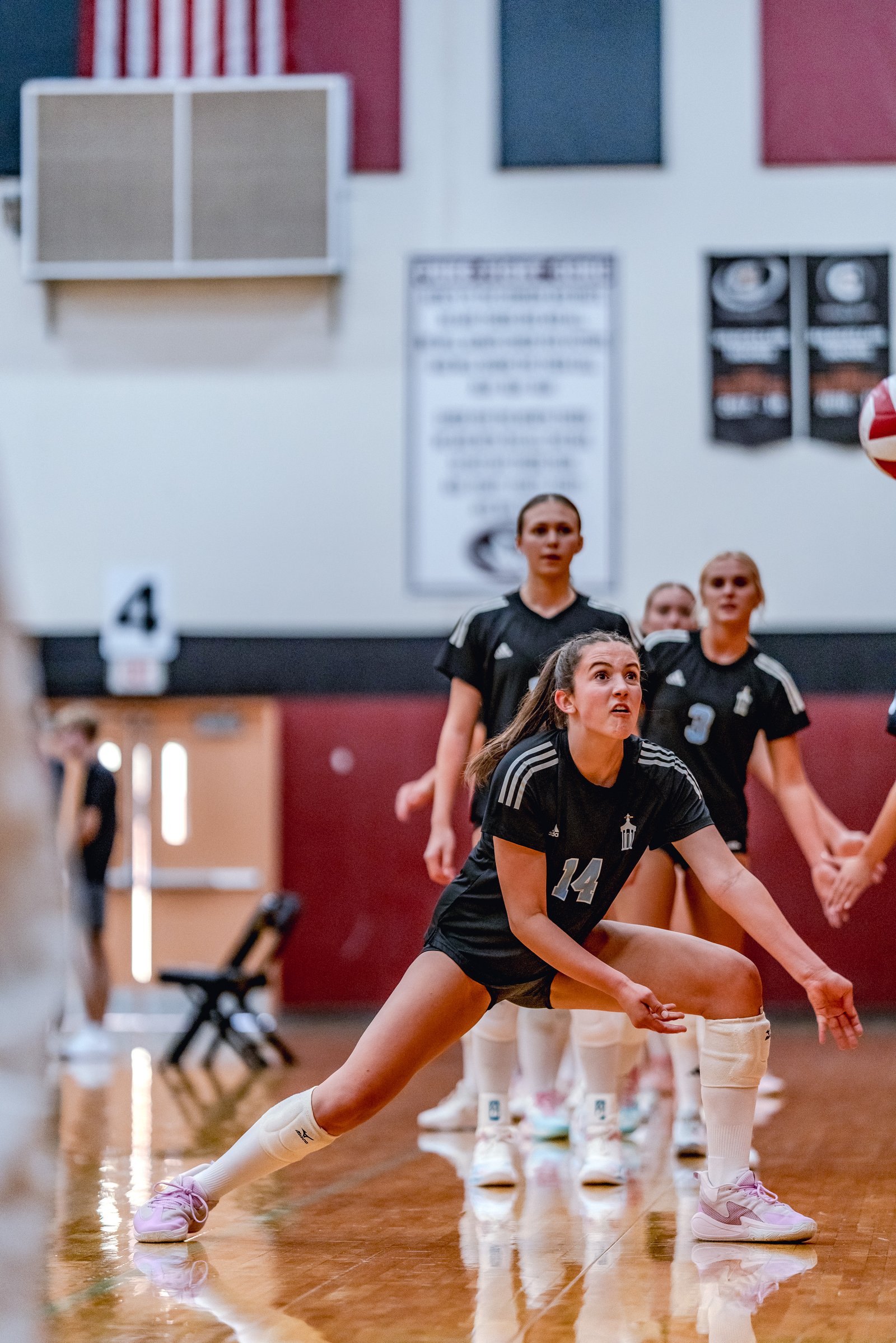A player in a black top with number 14 digs a volleyball on the court with teammates in black behind her.