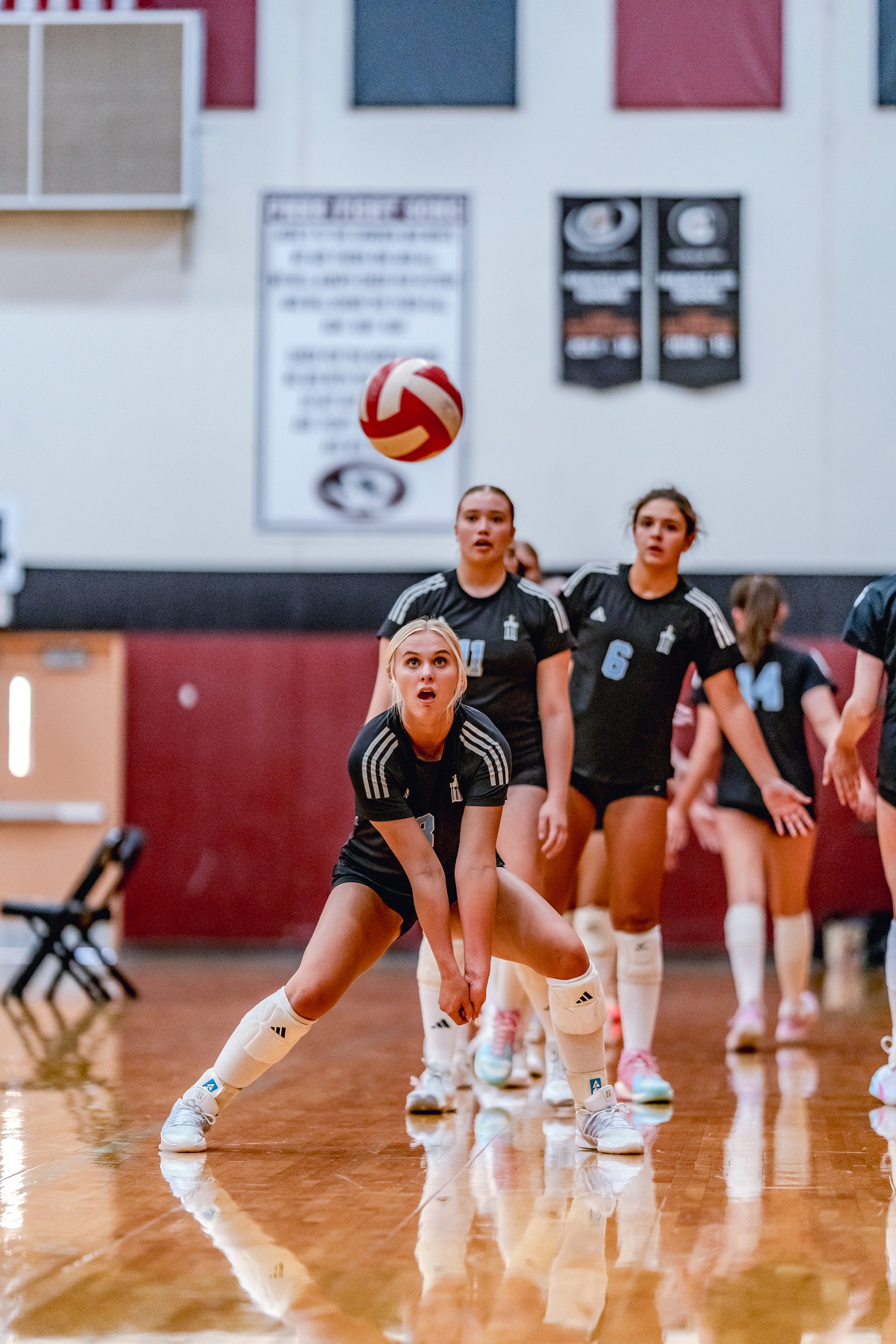 A player in black with white stripes prepares to dig a volleyball in a gym with other players in black uniforms.