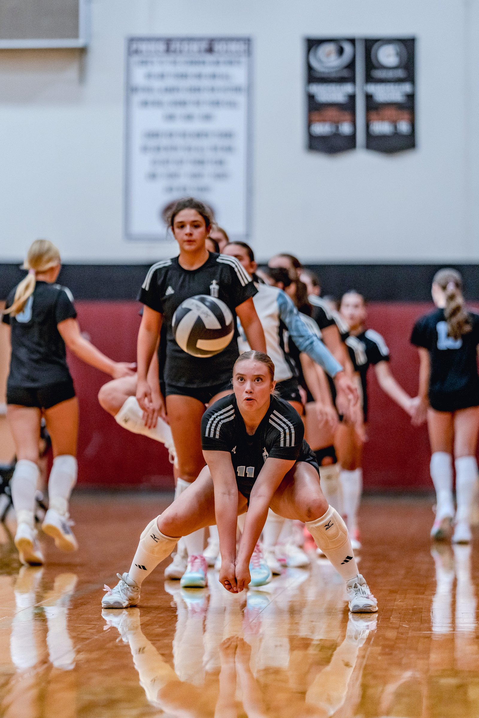 A player in black with white stripes crouches low, ready to receive a volleyball, with teammates in black and white uniforms behind her.
