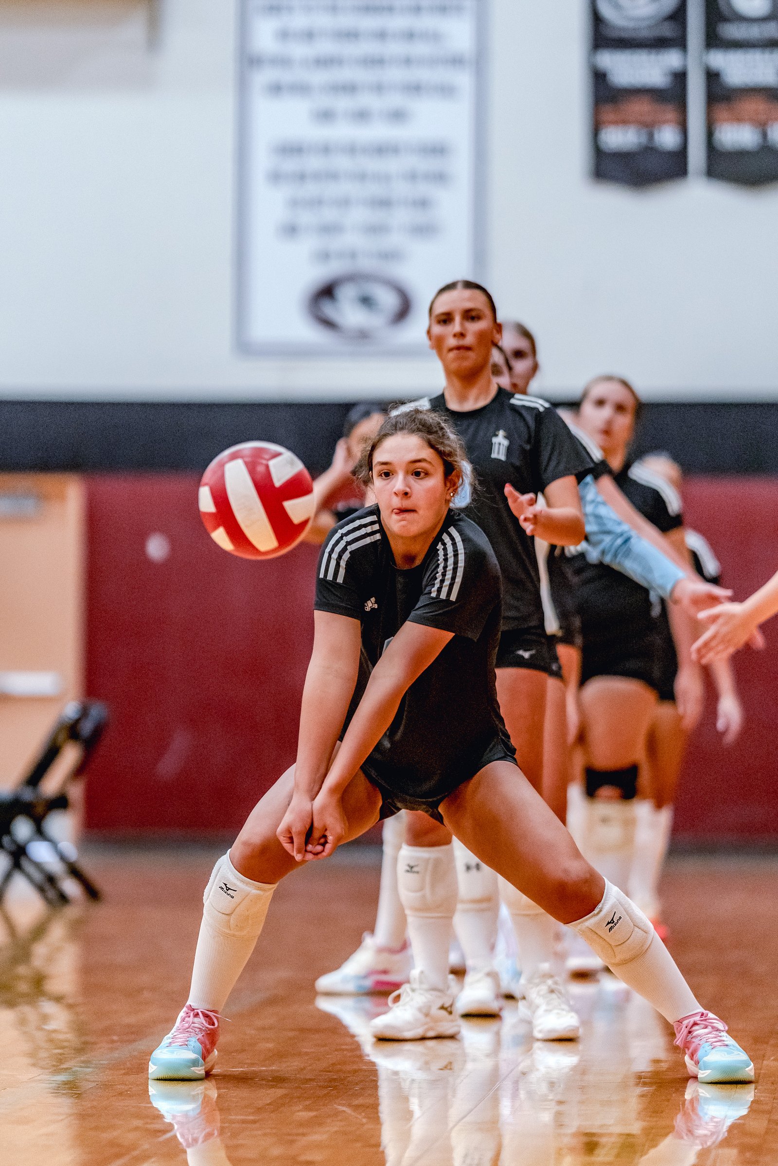 A player in black prepares to dig a volleyball as teammates in black and white uniforms line up behind her on the court.