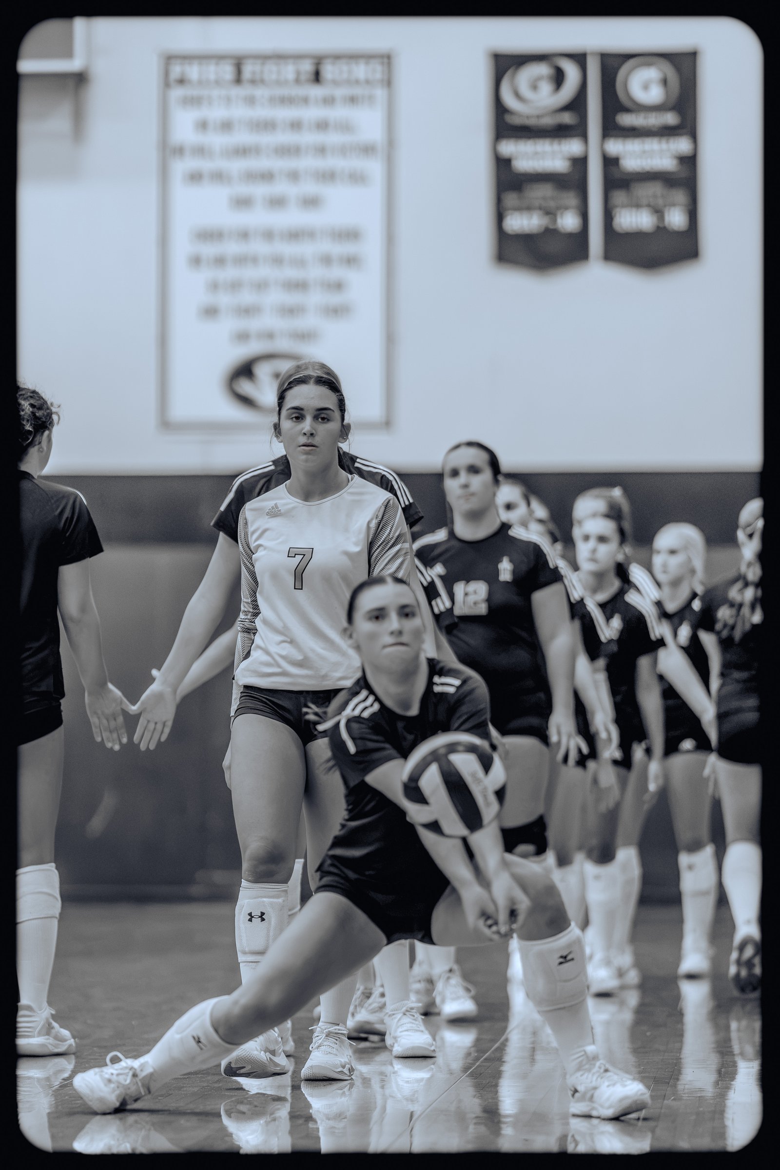 A player in a white jersey with number 7 attempts a dig as teammates in black jerseys watch during a volleyball game.