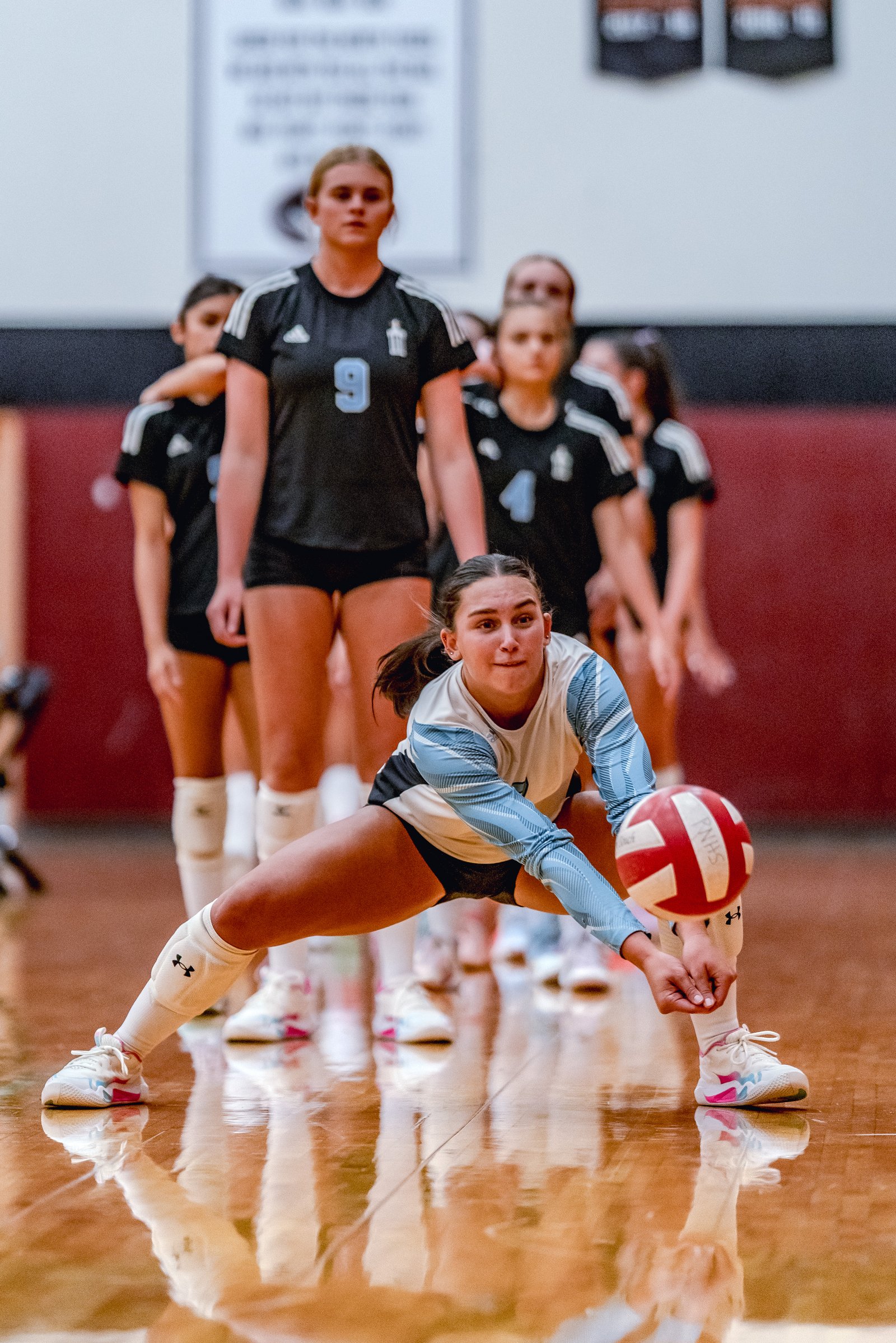 A player in a white and blue top dives for a volleyball, with teammates in black jerseys behind her.