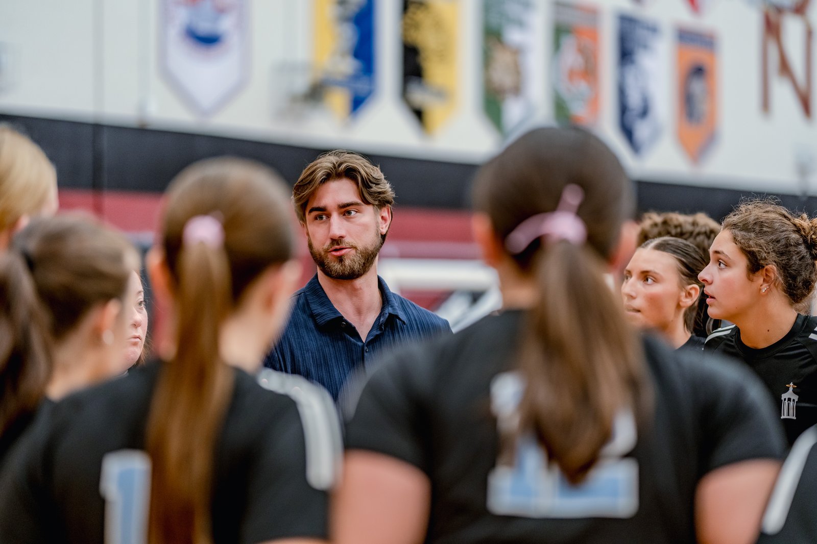 A coach talks to players in black jerseys with white trim during a volleyball game.