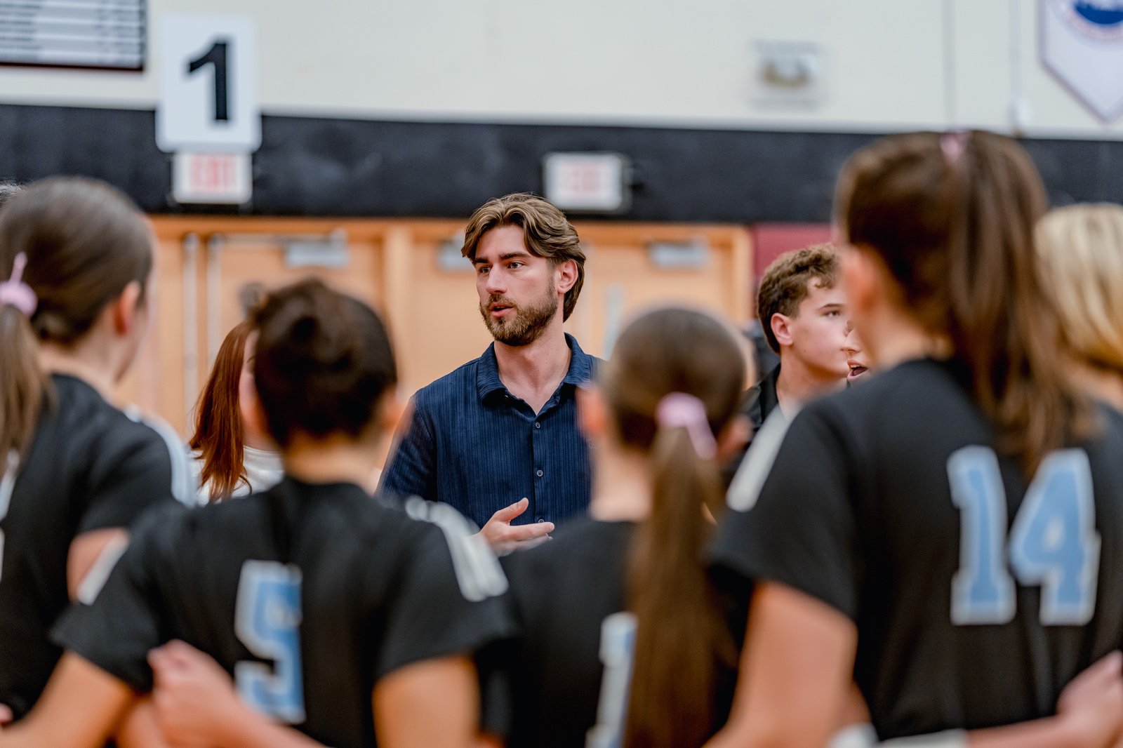 A coach speaks to players in black jerseys with light blue numbers, including 5 and 14, during a break in the game.